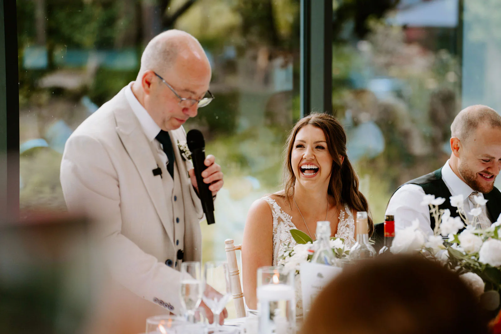 A man in a cream suit reads into a microphone beside the bride, who is laughing, and the groom, who is smiling, at the top table in a glass-sided room