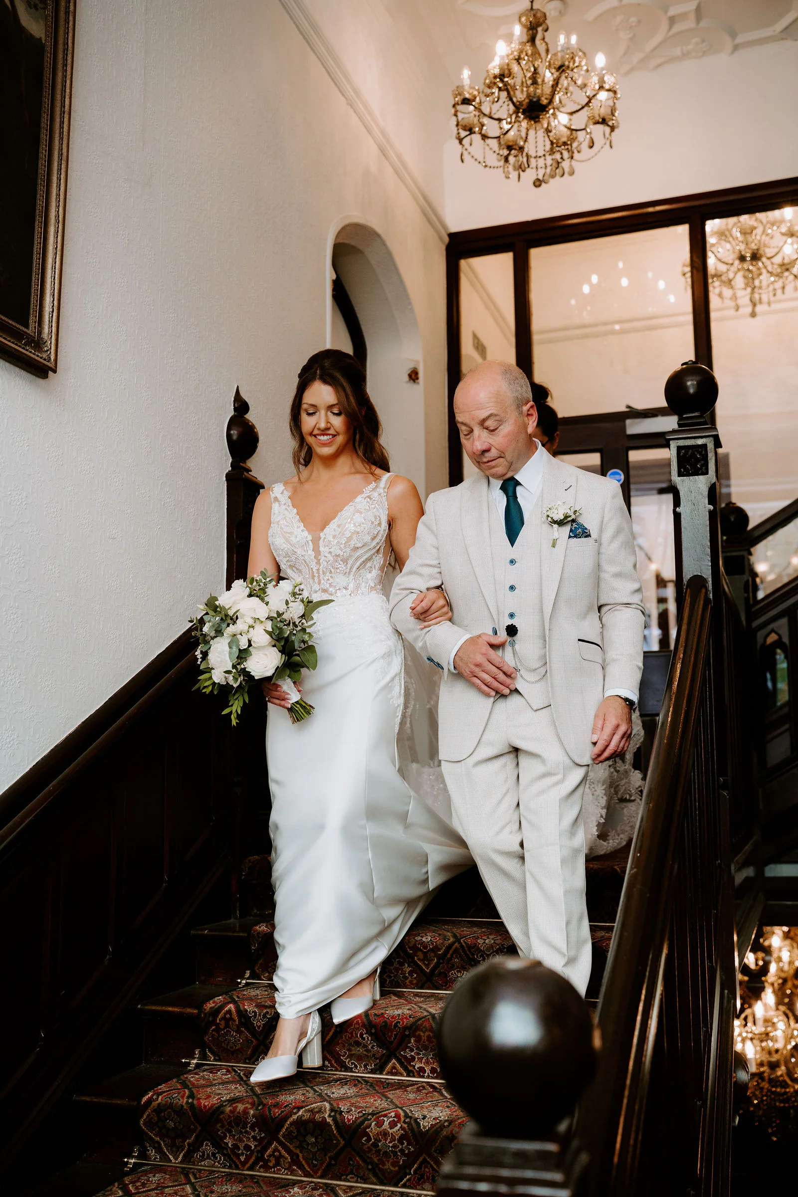 A bride holding a bouquet of white roses walks down a wooden staircase on the arm of her father in a cream suit, under a chandelier