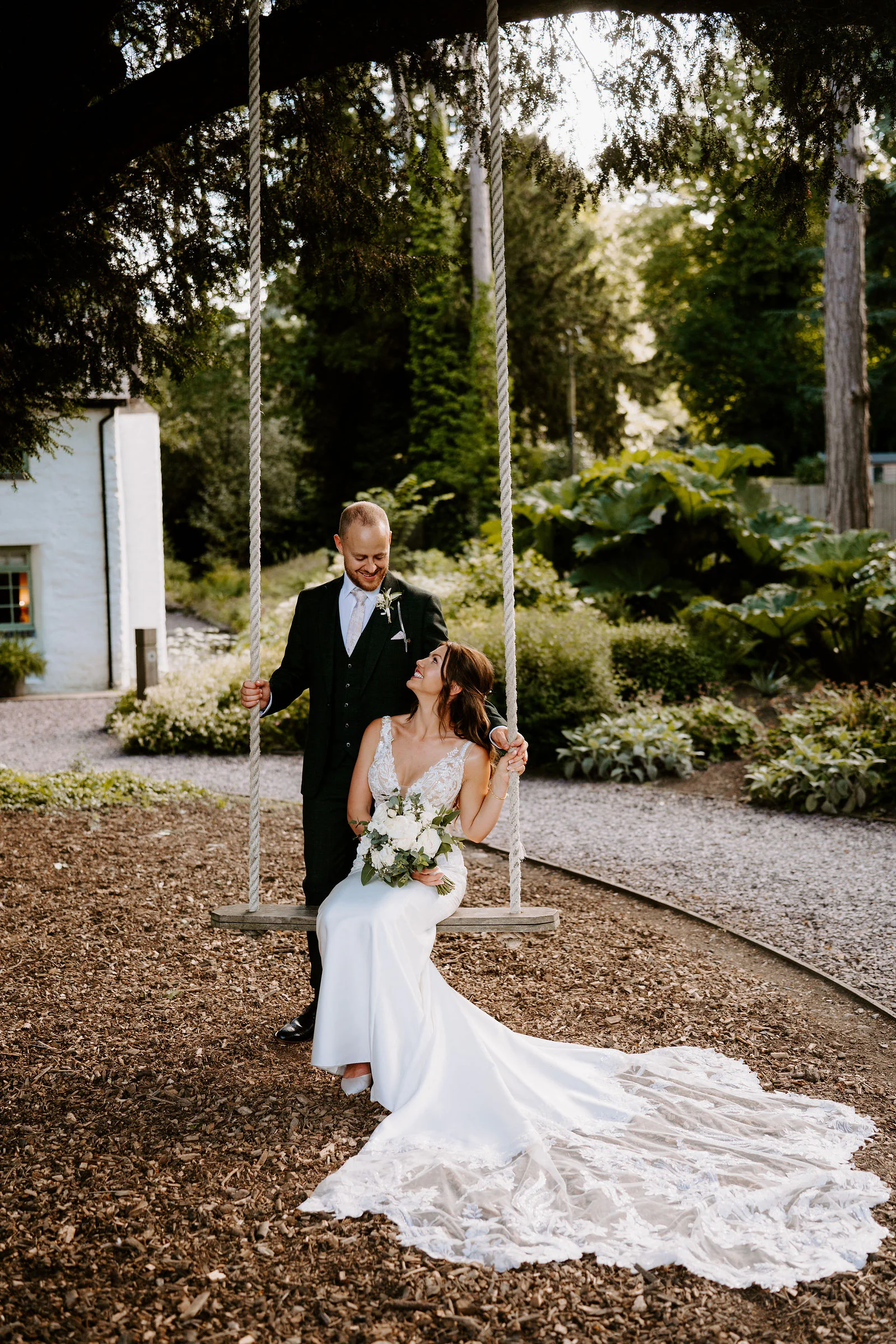 A bride sits on a rope swing hanging from a tree, her lace train spread across the bark chippings, while her groom in a dark suit stands behind her and smiles down at her, with a whitewashed building and tall trees beyond