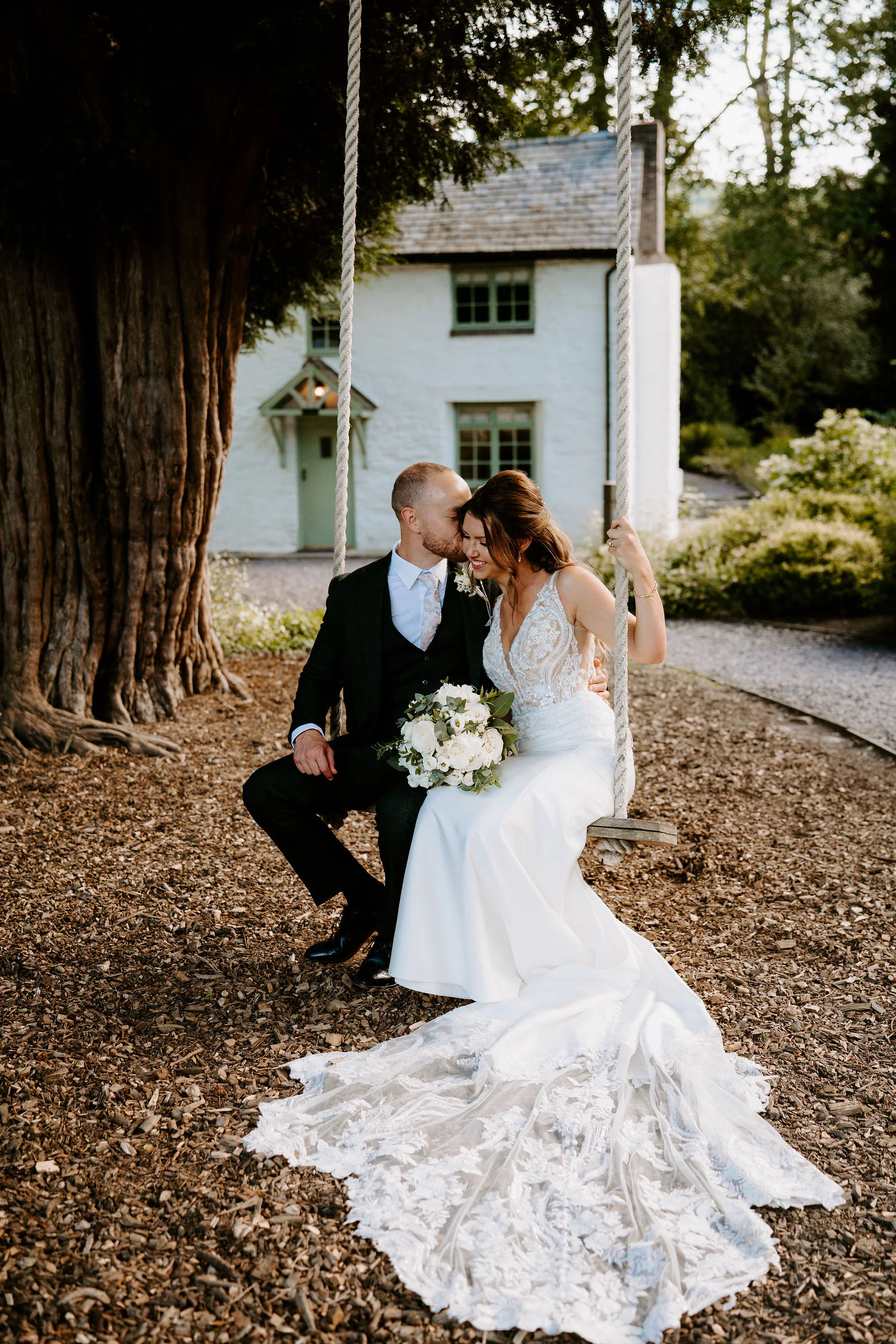 A bride in a lace gown with a very long train sits on a rope swing in the garden while her groom in a dark suit sits close beside her with his forehead against hers, a whitewashed cottage and a huge old tree trunk behind them at Tyn Dwr Hall
