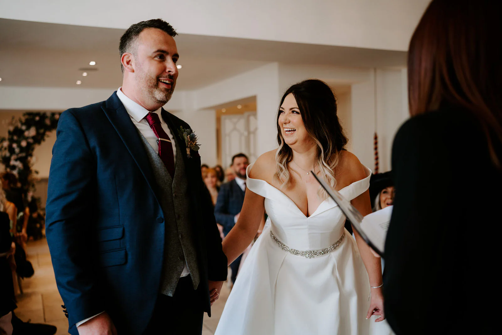 A groom in a navy suit and a smiling bride in an off-the-shoulder gown walk together along the aisle, a woman in black beside them and seated guests beyond