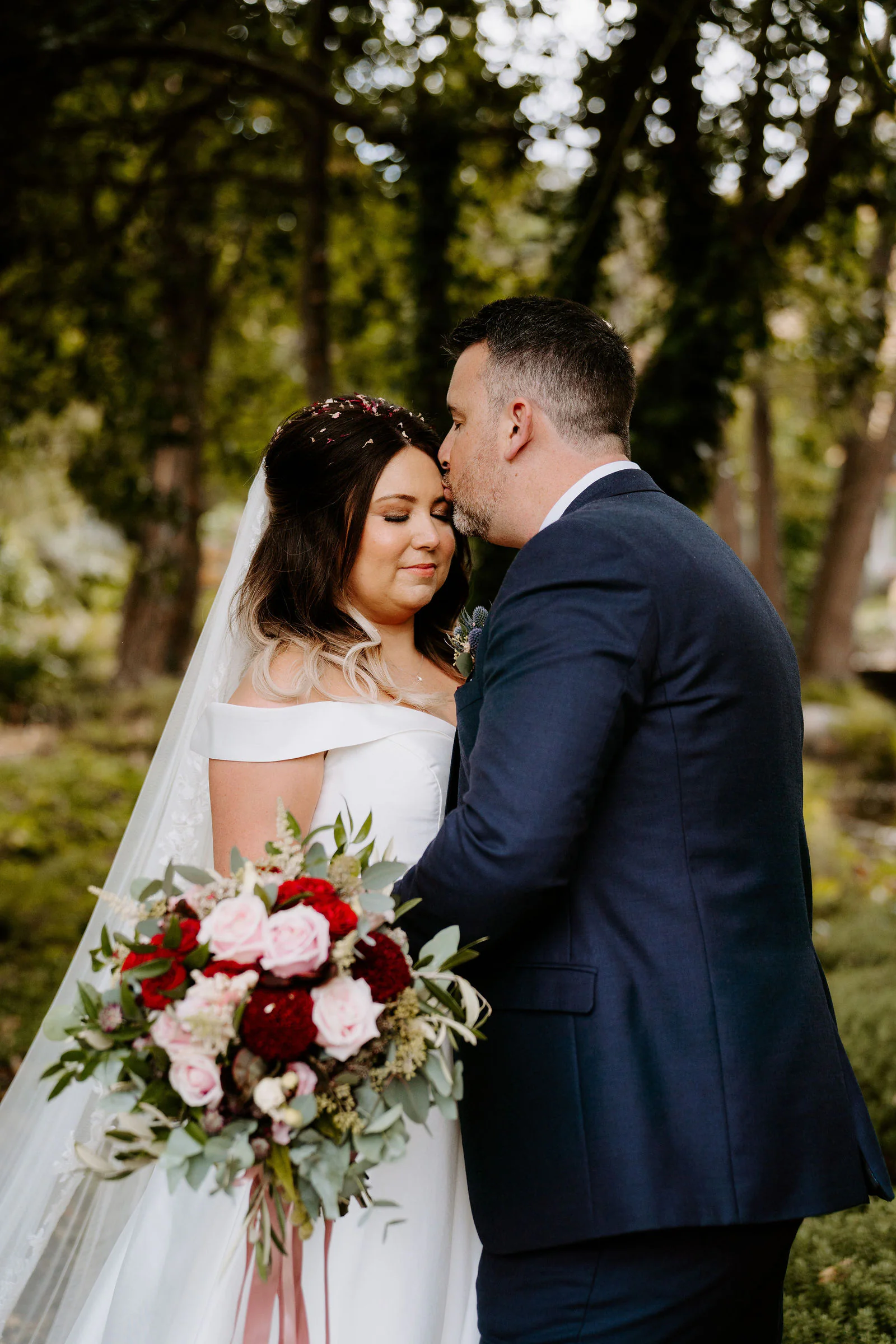 A groom in a navy suit kisses the cheek of a bride in a veil and off-the-shoulder gown who holds a bouquet of red and pink roses, with trees behind them
