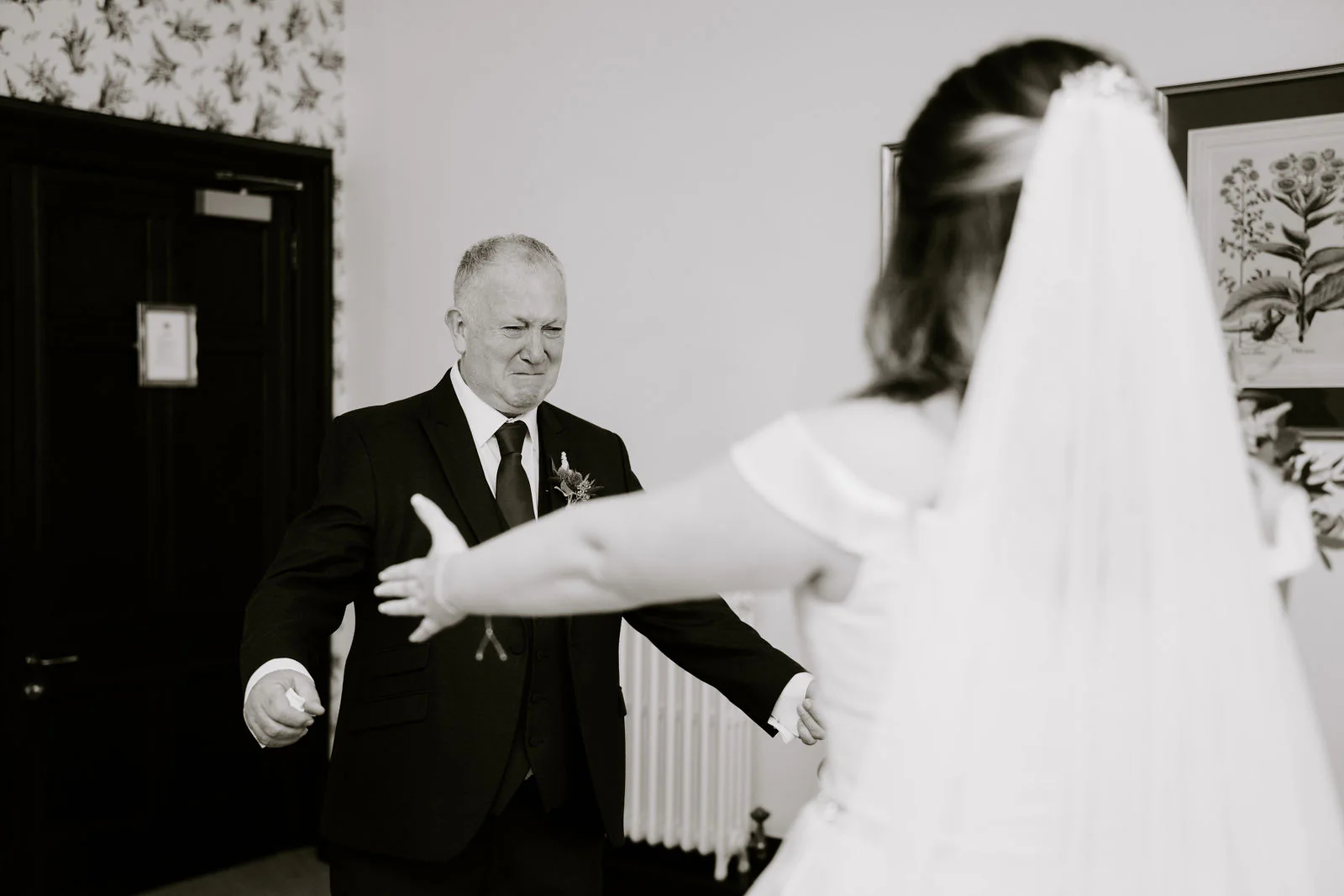 In black and white, a man in a dark suit opens his arms to hug a bride in a veil who reaches towards him, in a room with patterned wallpaper