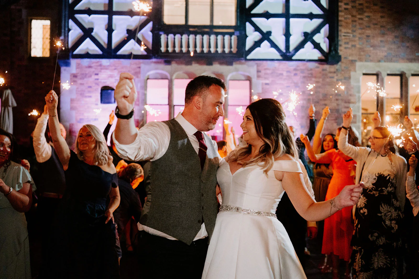 A groom in a tweed waistcoat and a bride in an off-the-shoulder gown dance with their guests as sparklers burn around them, the black-and-white timbered front of the hall lit up behind