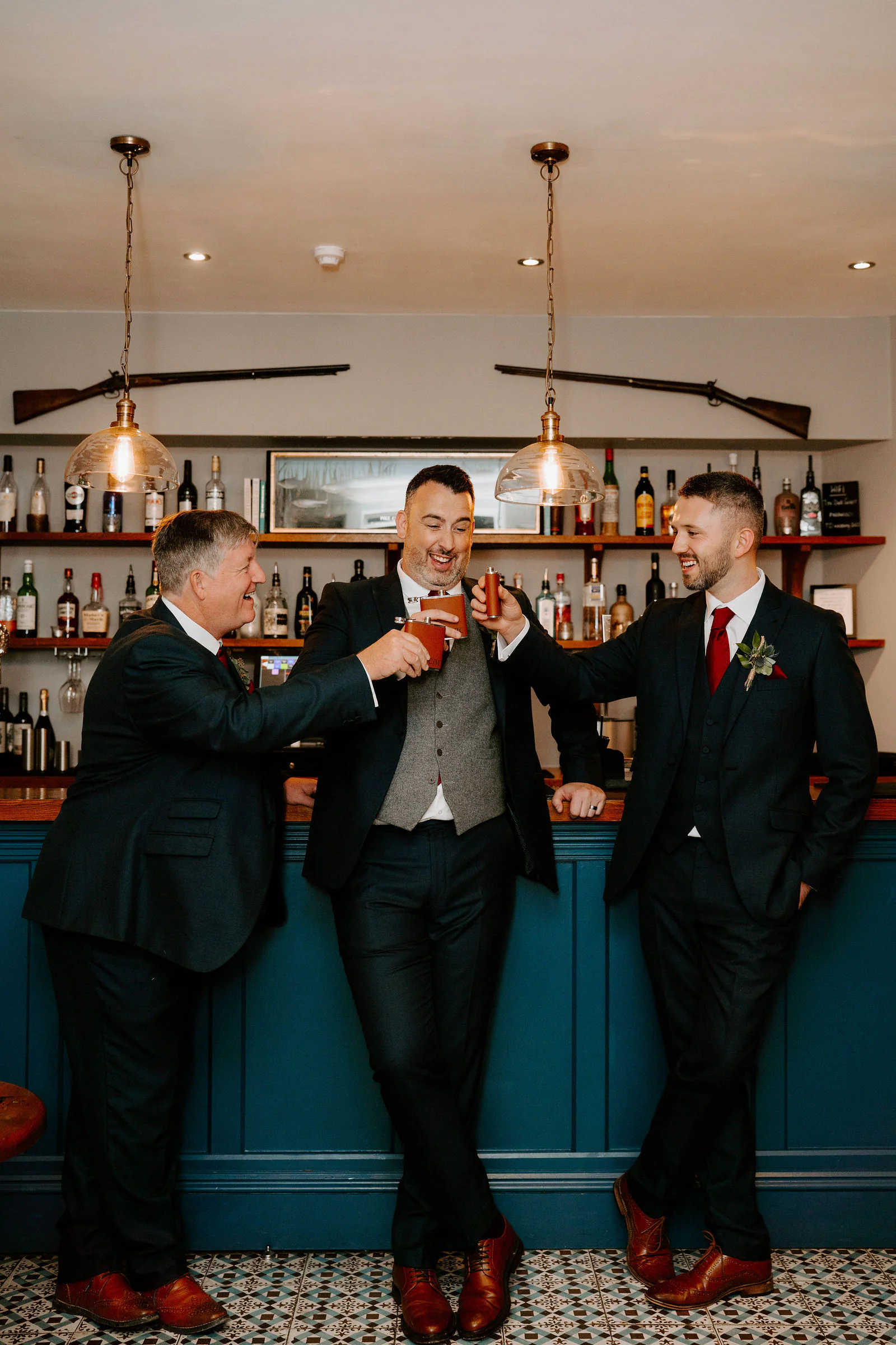 Three men in dark suits and waistcoats raise glasses in a toast, leaning against a teal bar with bottles and two hanging lamps behind them