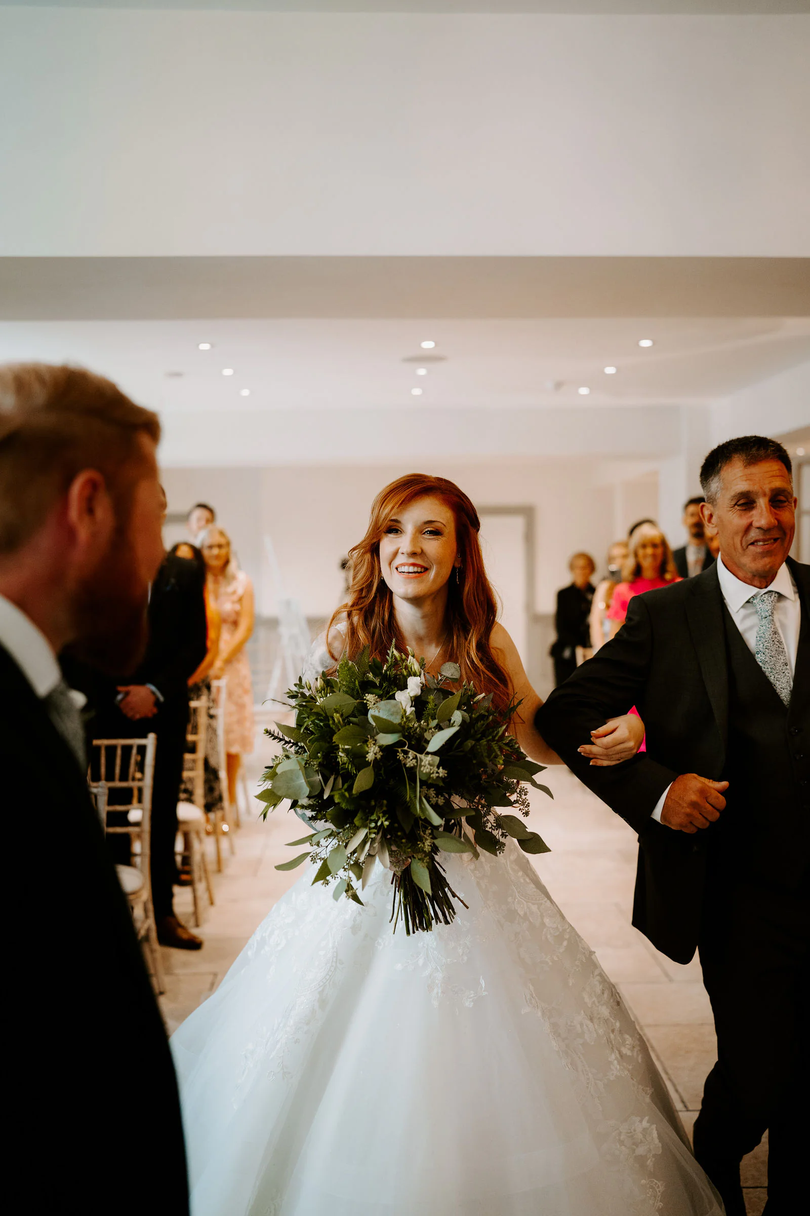 A bride in a ball gown with a green bouquet walks up the aisle smiling, on the arm of the man beside her, past the groom’s head in the foreground and guests beyond