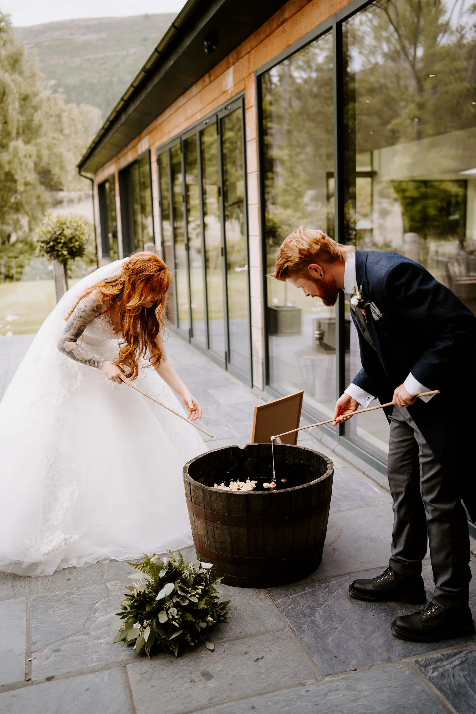 A bride and groom bend over an oak barrel holding long sticks outside a glass-fronted building, a wreath of foliage on the slate floor beside them and wooded hills behind