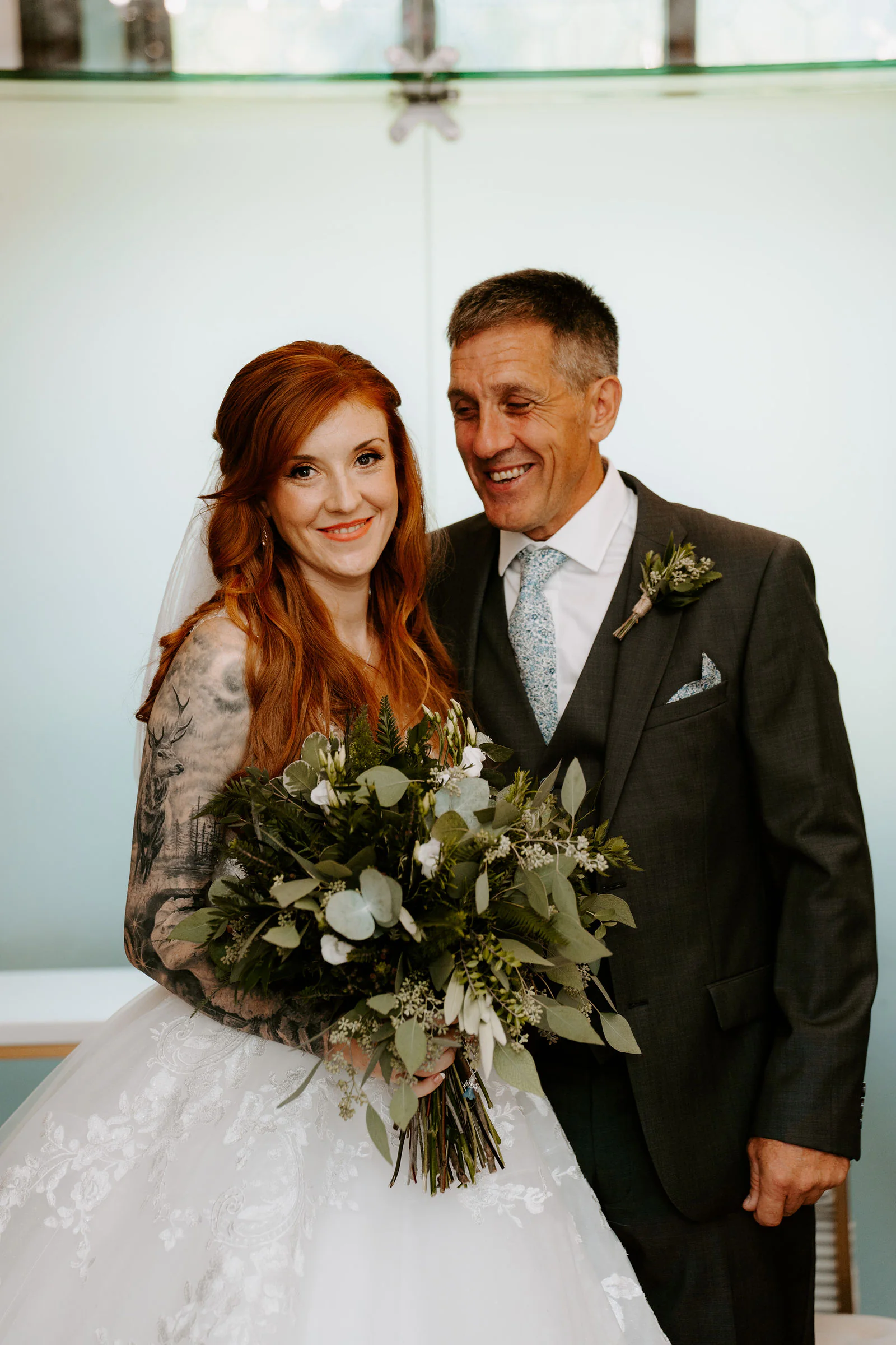 A bride with long red hair and a tattooed arm holds a bouquet of green foliage and smiles beside her father, who is laughing, in a bright room