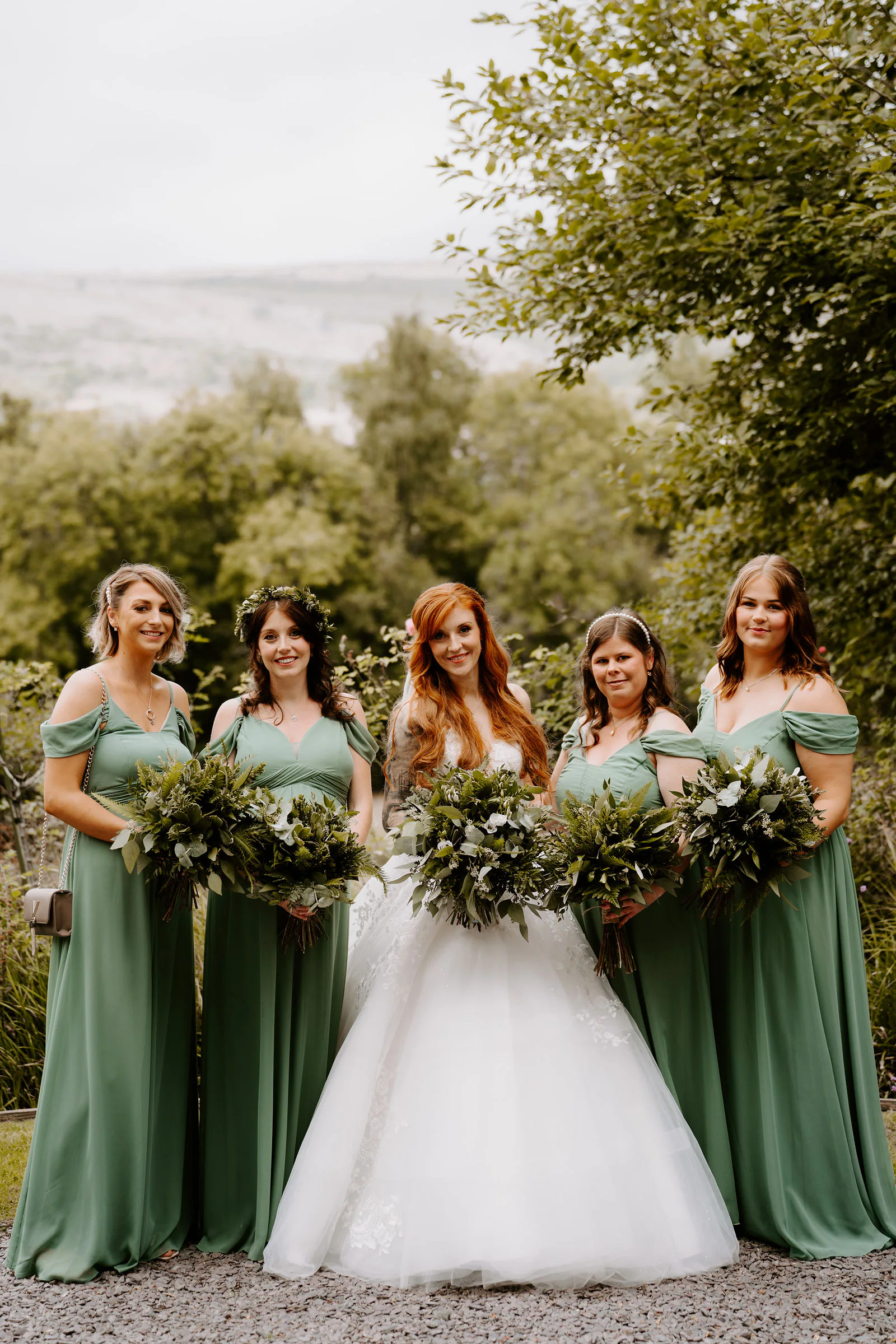 A bride in a ball gown stands with five bridesmaids in sage green dresses holding bouquets of foliage on a gravel path, with trees and hills behind them