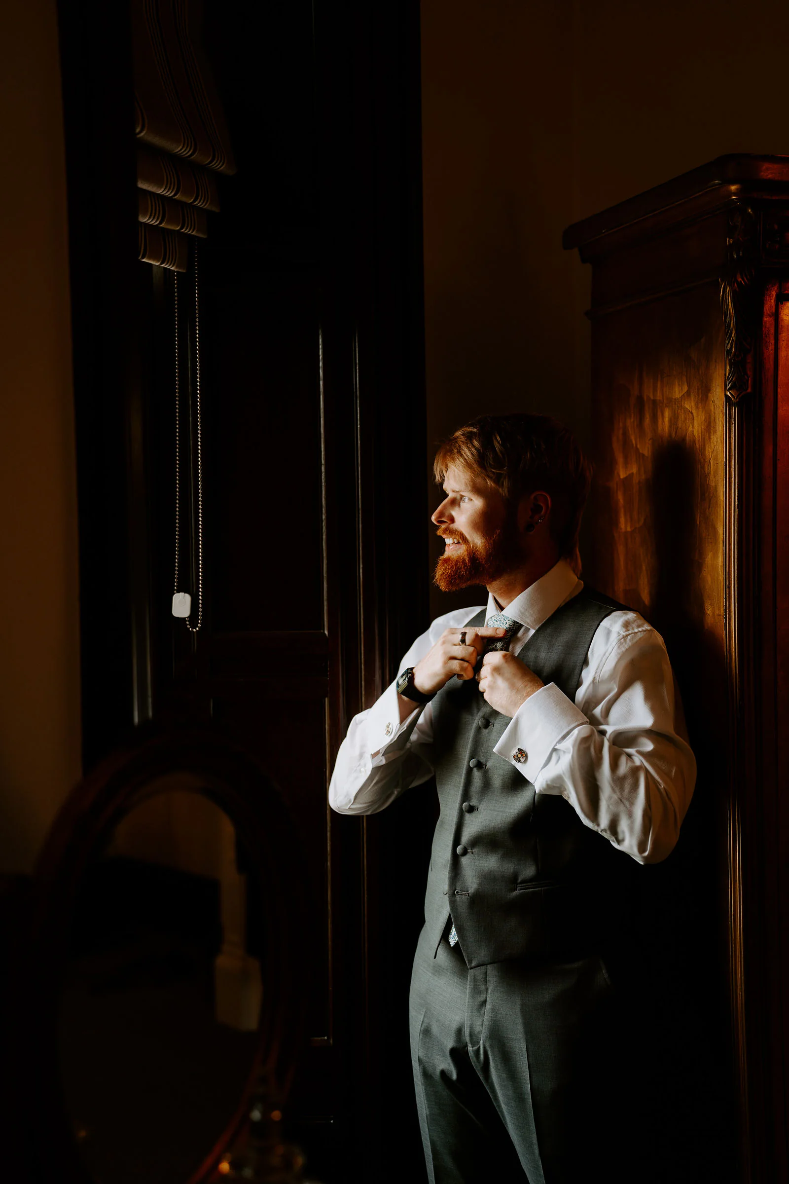 A groom with a red beard adjusts the collar of his waistcoat in the shadow of a dark panelled room, looking out of a window beside a wardrobe