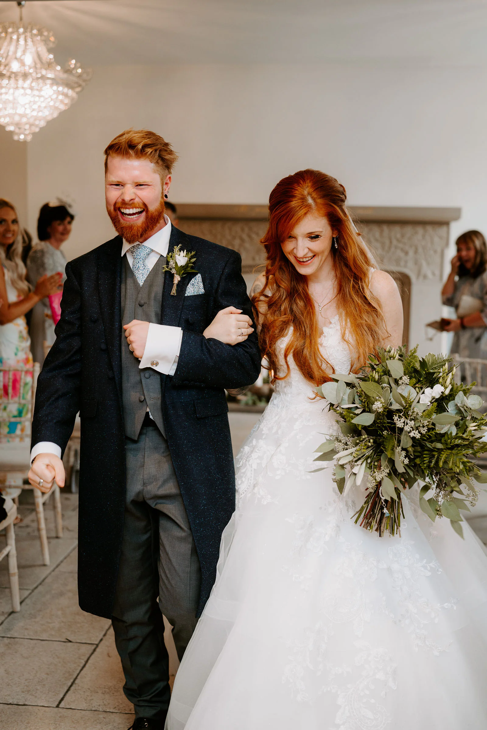 A groom in a navy coat and a bride in a lace gown walk back down the aisle together, both laughing, her arm through his, with guests behind them