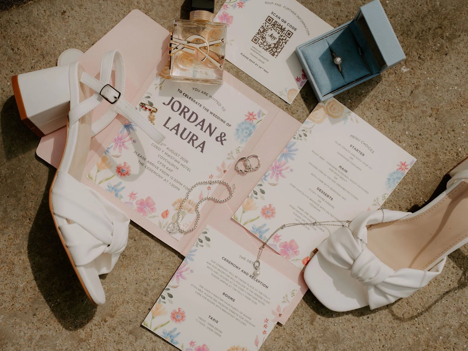 A flat lay on stone of a wedding invitation reading Jordan and Laura, white block-heeled sandals, a bottle of perfume, a blue ring box, pearl jewellery and printed menus