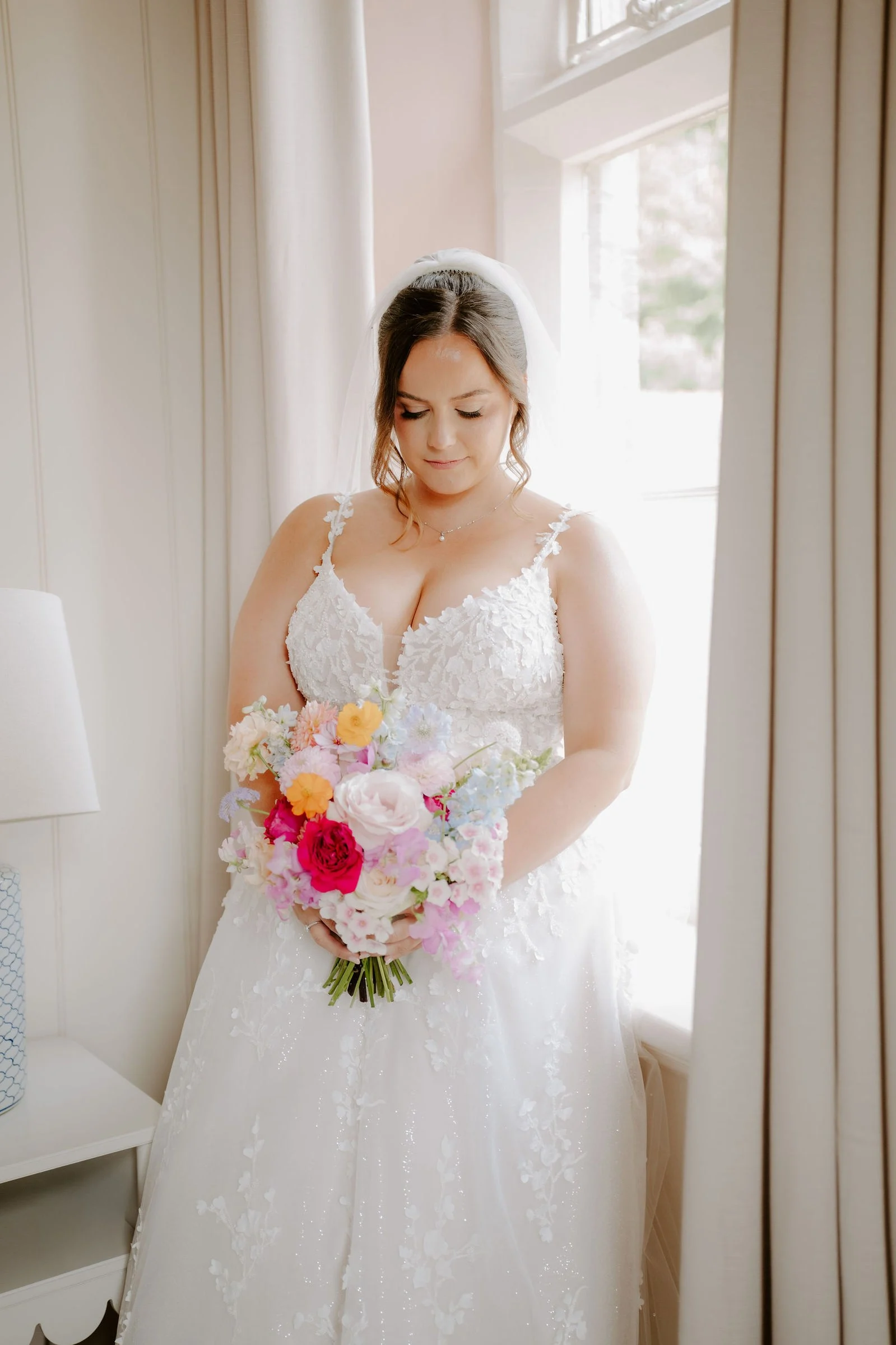 A bride in a lace wedding dress and veil stands beside a tall window holding a bright bouquet of pink, orange and white flowers, looking down.