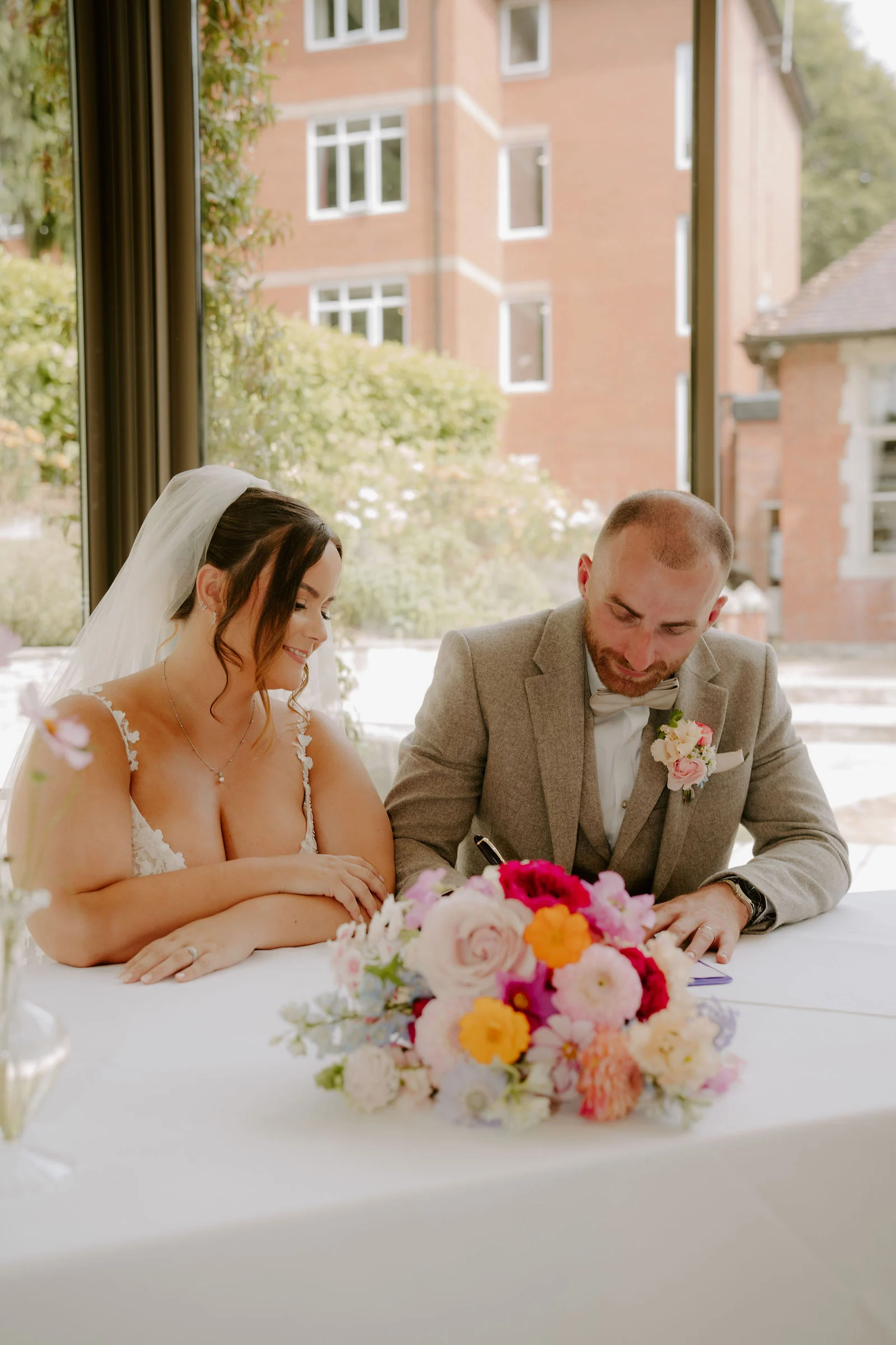 A bride in a veil and a groom in a beige jacket sign the register at a table with a bouquet of bright flowers, a glass wall and a red-brick building behind them