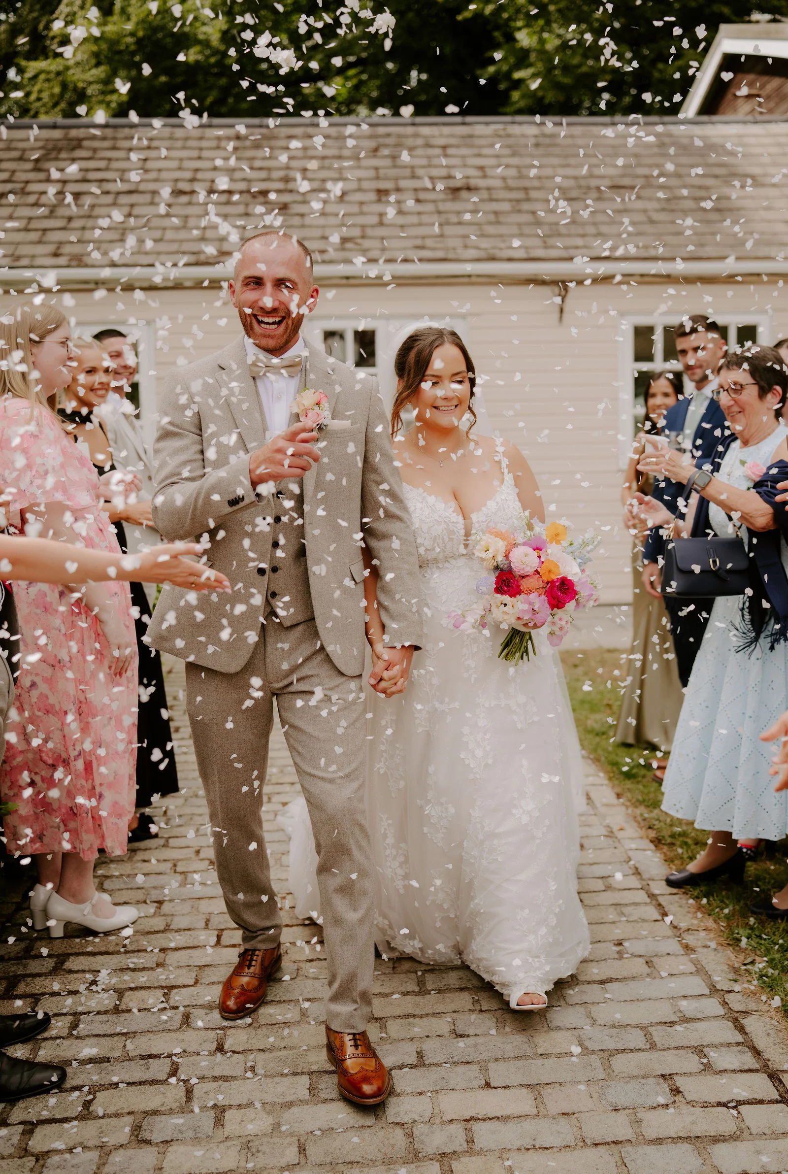 A laughing groom and a bride holding a bouquet walk hand in hand down a cobbled path while guests throw white confetti around them.