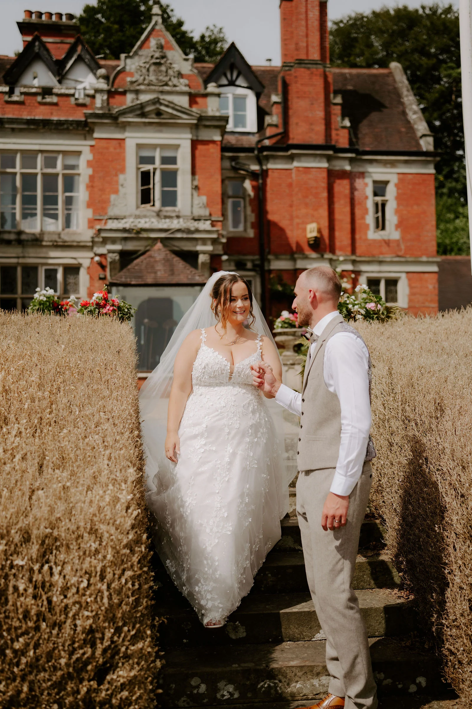 A bride in a long veil and lace dress walks down garden steps between tall golden hedges towards a groom in a waistcoat, with a red brick manor house behind them.