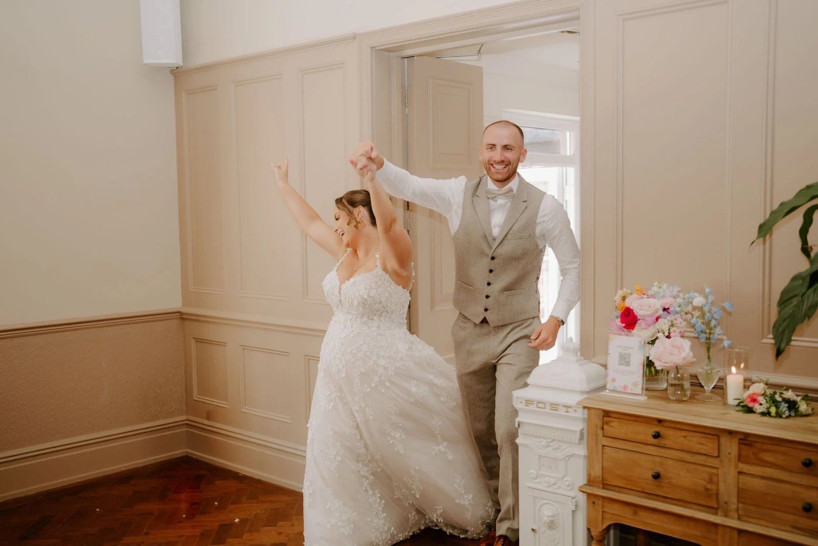 A bride in a lace gown and a groom in a waistcoat walk into a cream panelled room with their arms raised in celebration, a wooden dresser with flowers behind them