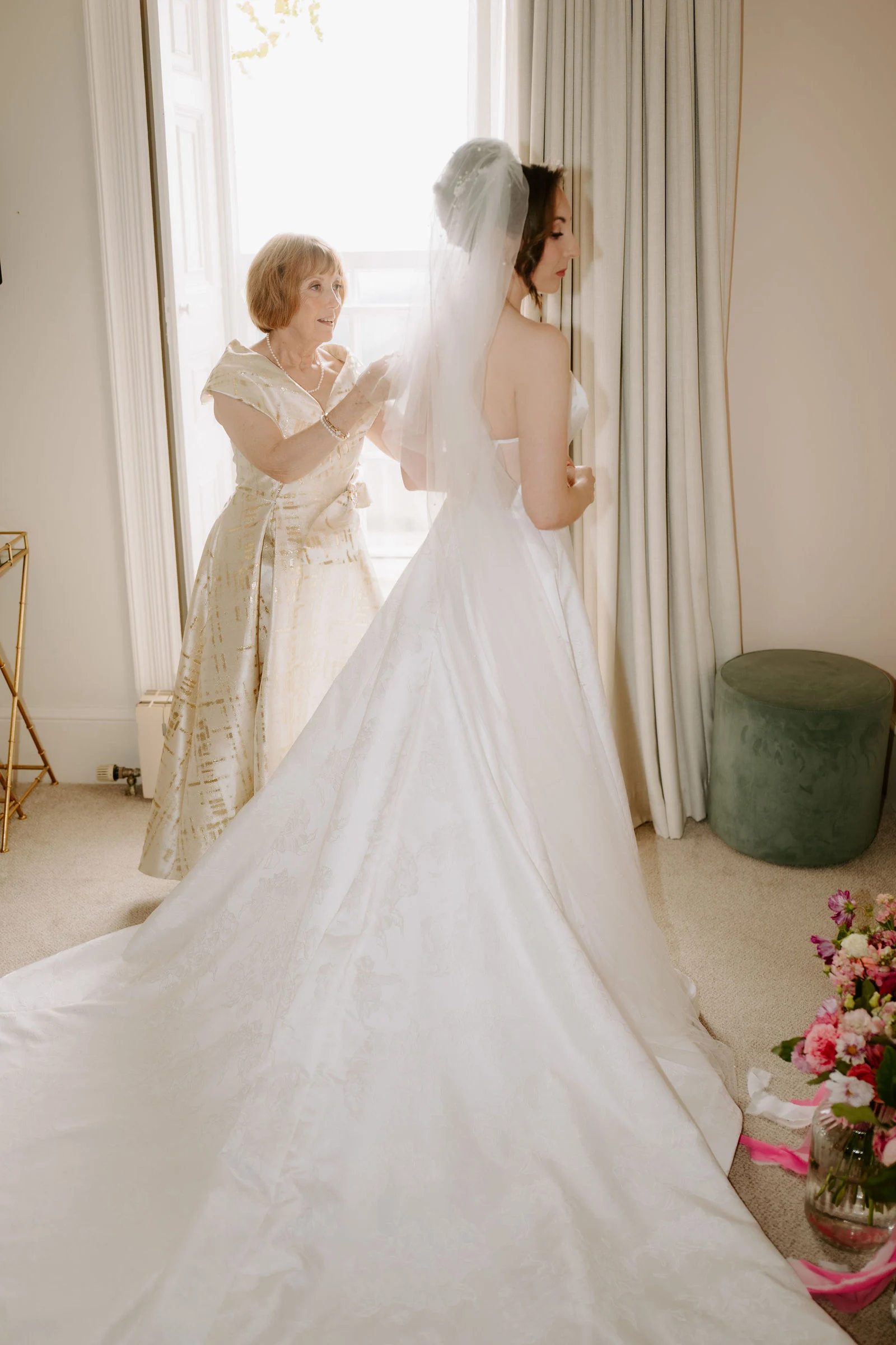 A bride in a strapless gown with a long train stands at a tall sunlit window while a woman in a gold patterned dress adjusts her veil