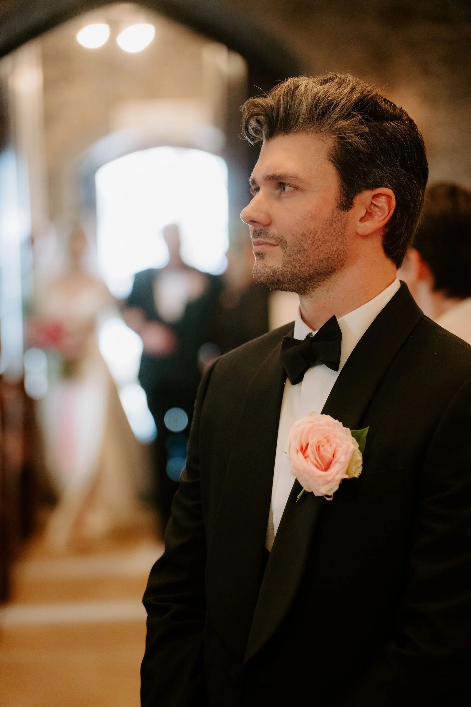 A groom in a black dinner jacket with a pink rose buttonhole looks down the aisle of a stone church while the bride walks in, blurred, behind him