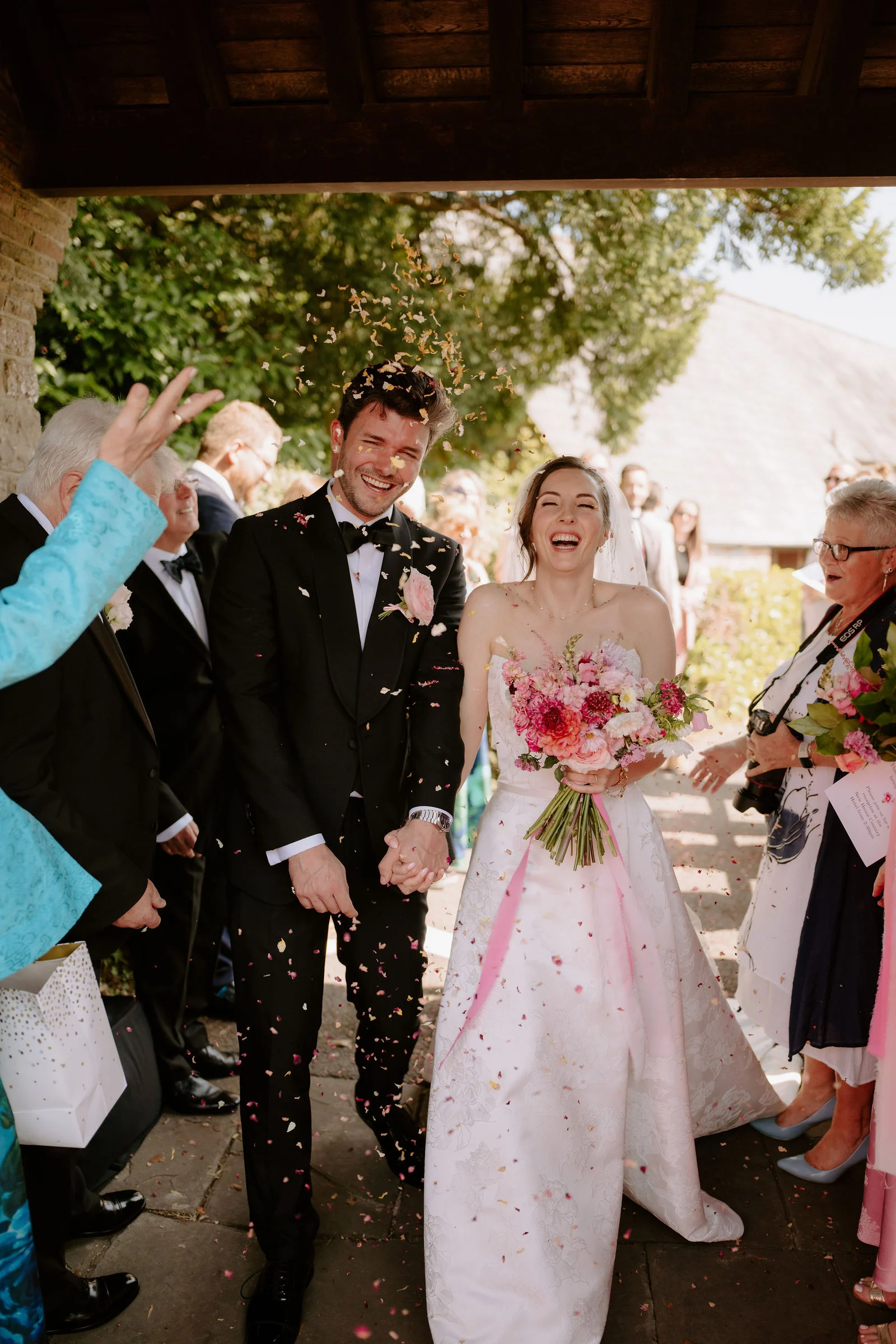 A newly married couple laugh as confetti falls around them under a church porch while guests cheer either side