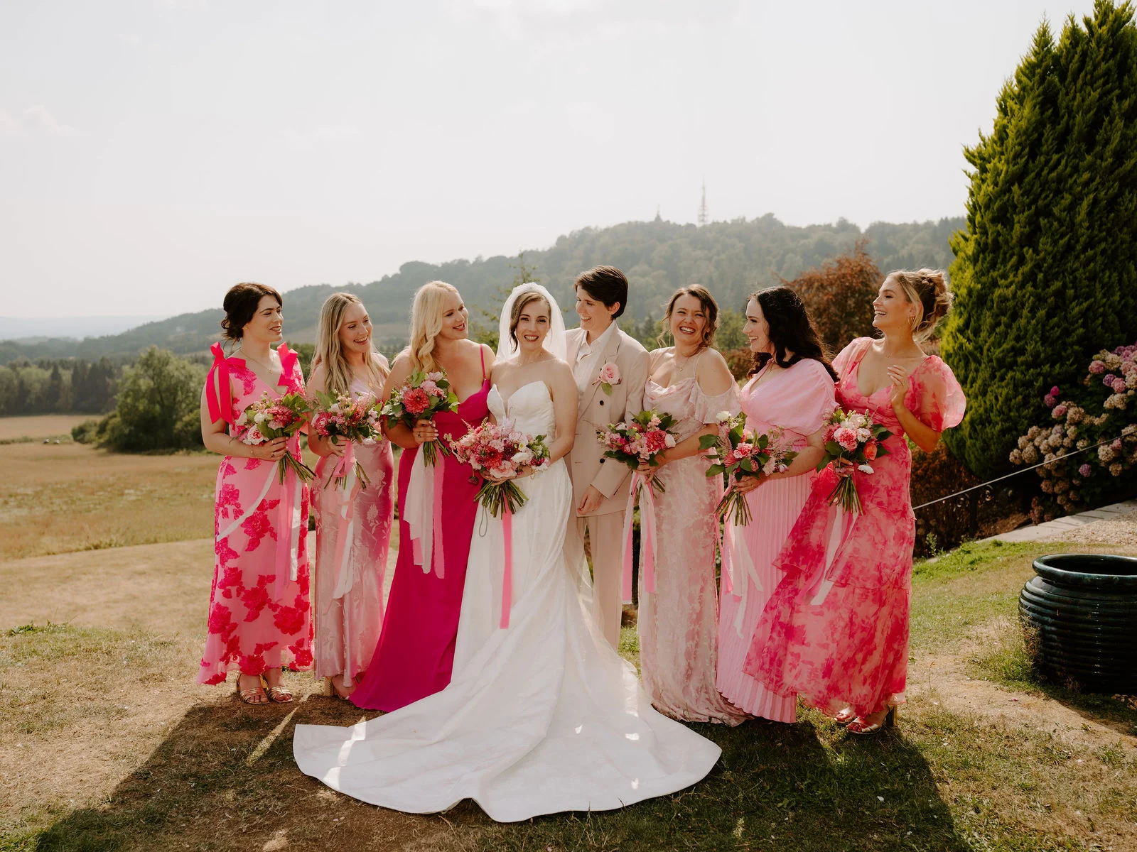A bride in a fitted white dress laughs with her bridesmaids in shades of pink on a lawn with wooded hills behind them