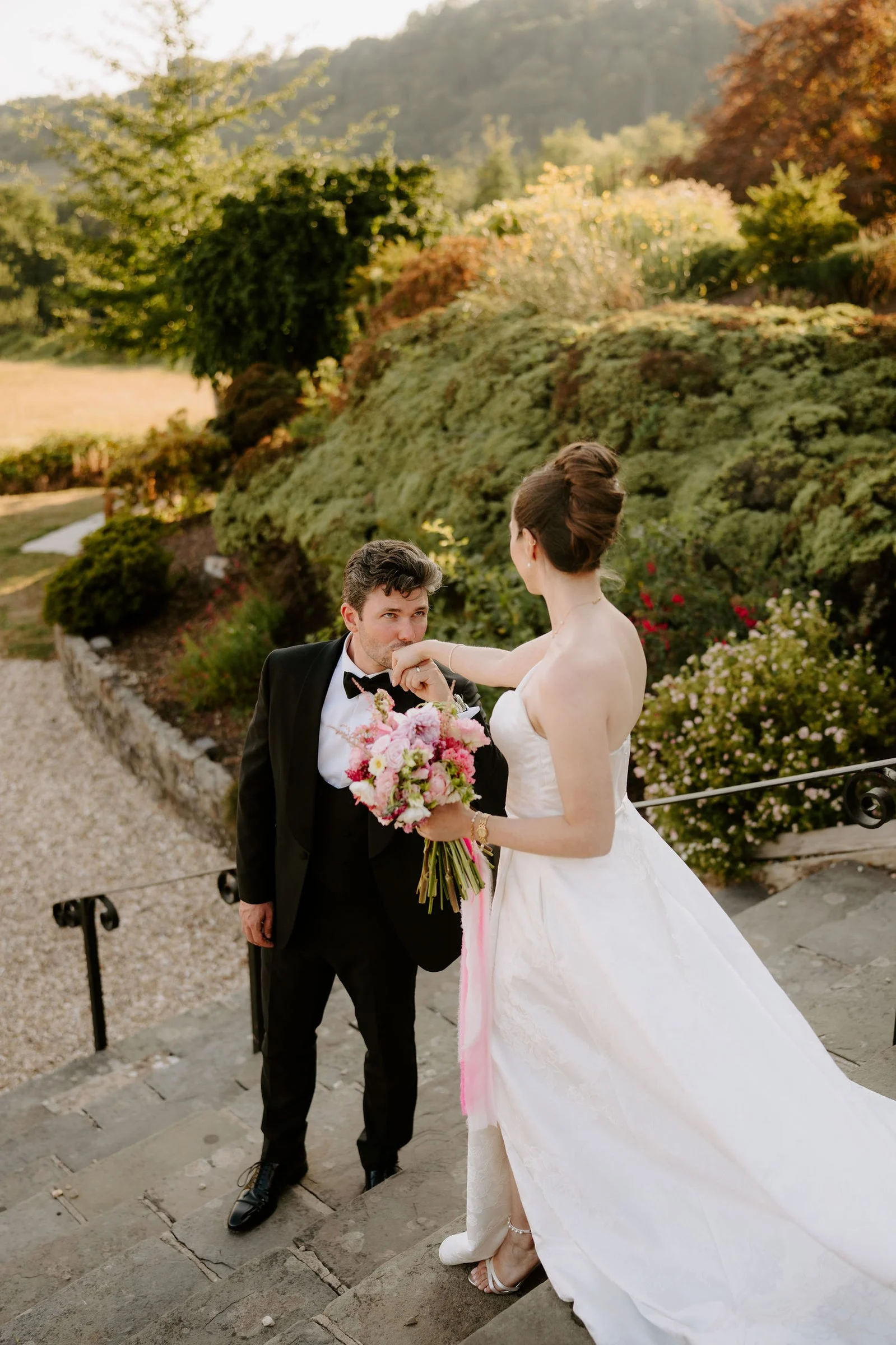 On a stone staircase in the hotel gardens a groom in a black suit kisses his bride's hand while she holds a pink bouquet