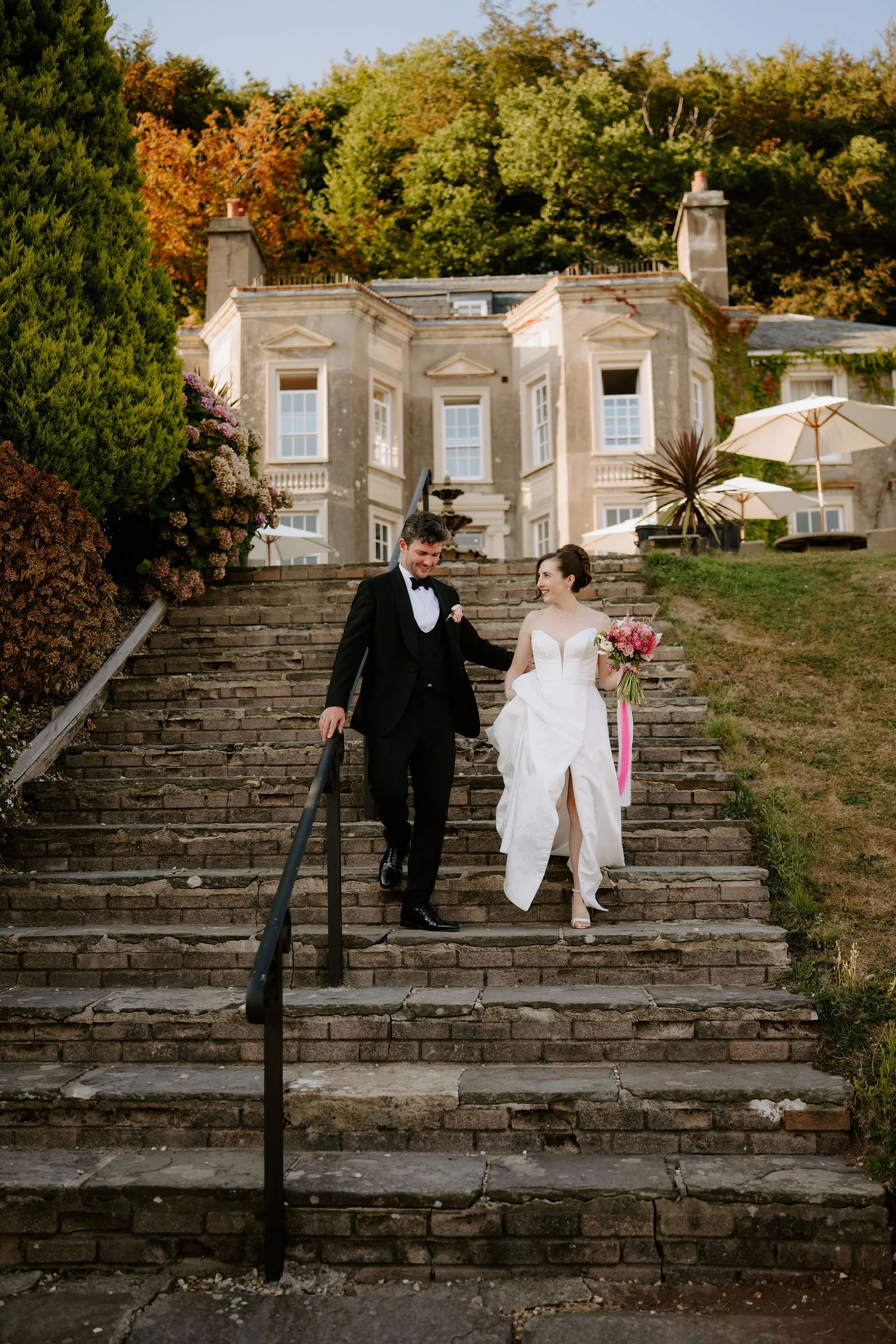 A couple in a white wedding dress and black suit walk hand in hand down a long flight of old stone steps with the manor house above them