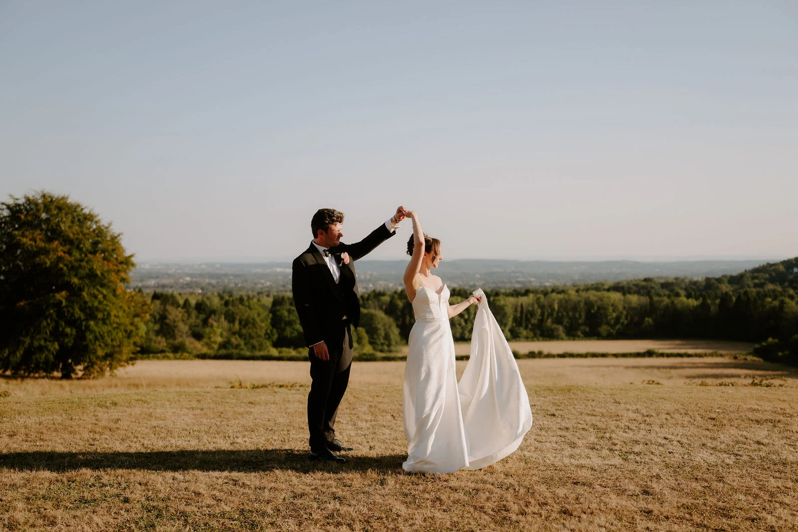 A groom twirls his bride on a dry golden lawn with a wide view of distant woodland and countryside in evening light
