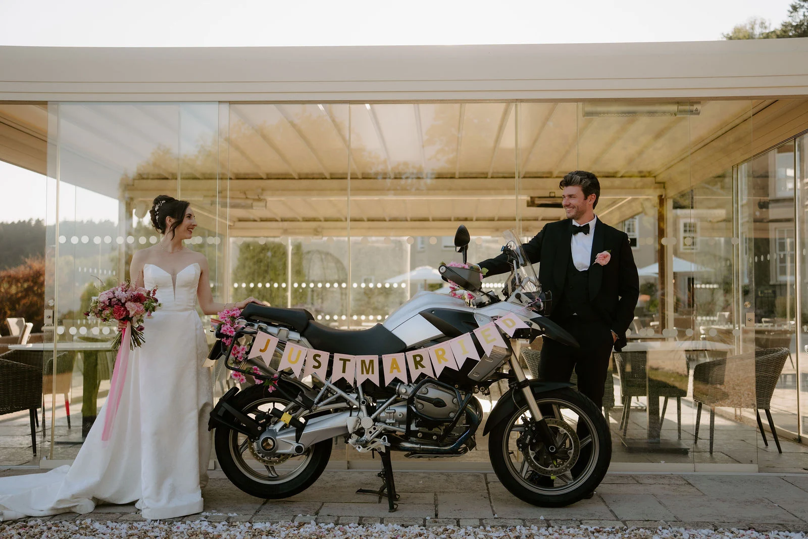 A bride and groom stand either side of a motorbike decorated with a Just Married banner under a glass-sided terrace