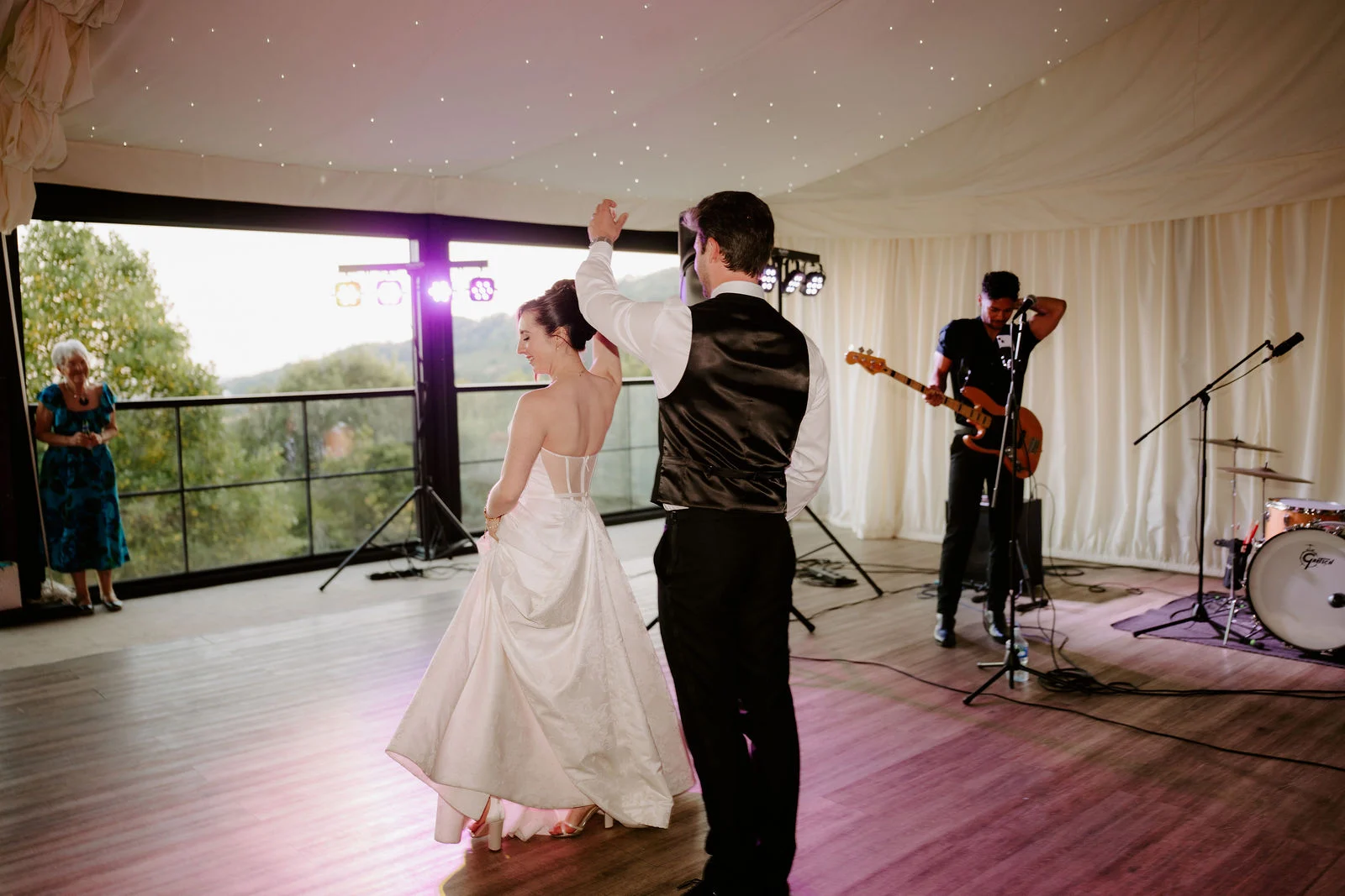 A bride and groom dance in a room with big windows and a canopy of fairy lights while a live band plays behind them in a purple glow