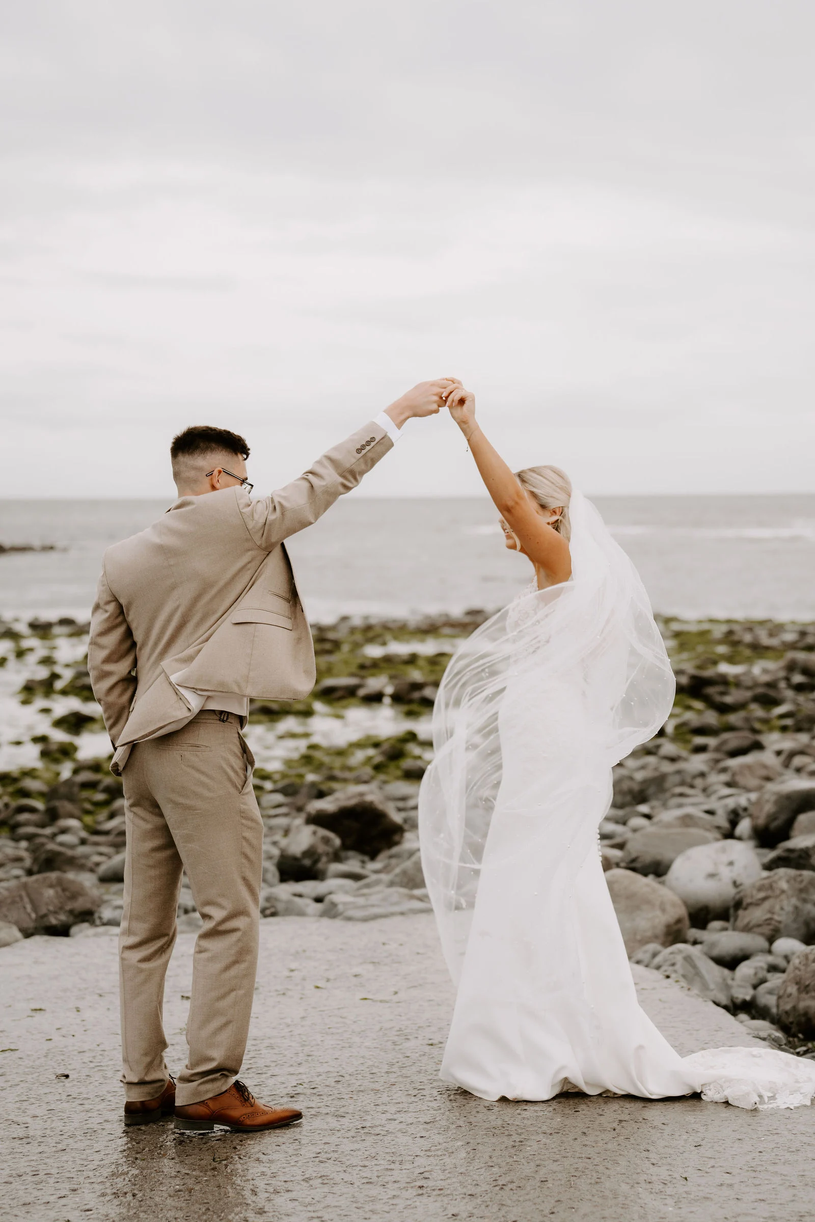 A groom in a tan suit lifts a bride's hand to twirl her, her long veil lifting in the wind, on a wet concrete path beside a rocky shore and grey sea.