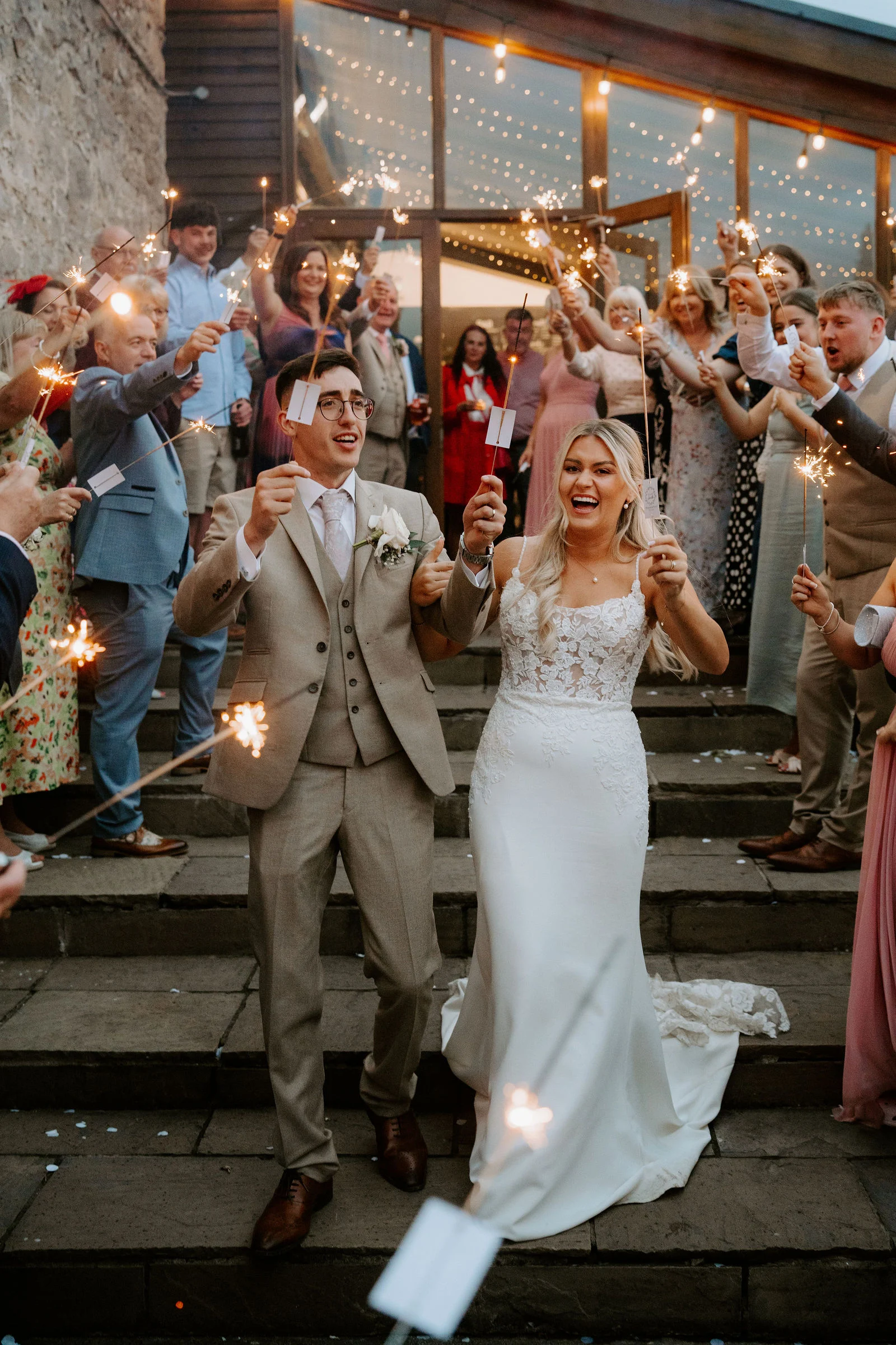 A groom in a beige suit and a bride in a fitted gown laugh as they come down stone steps through guests holding sparklers on both sides, with a glass-fronted building hung with lights behind