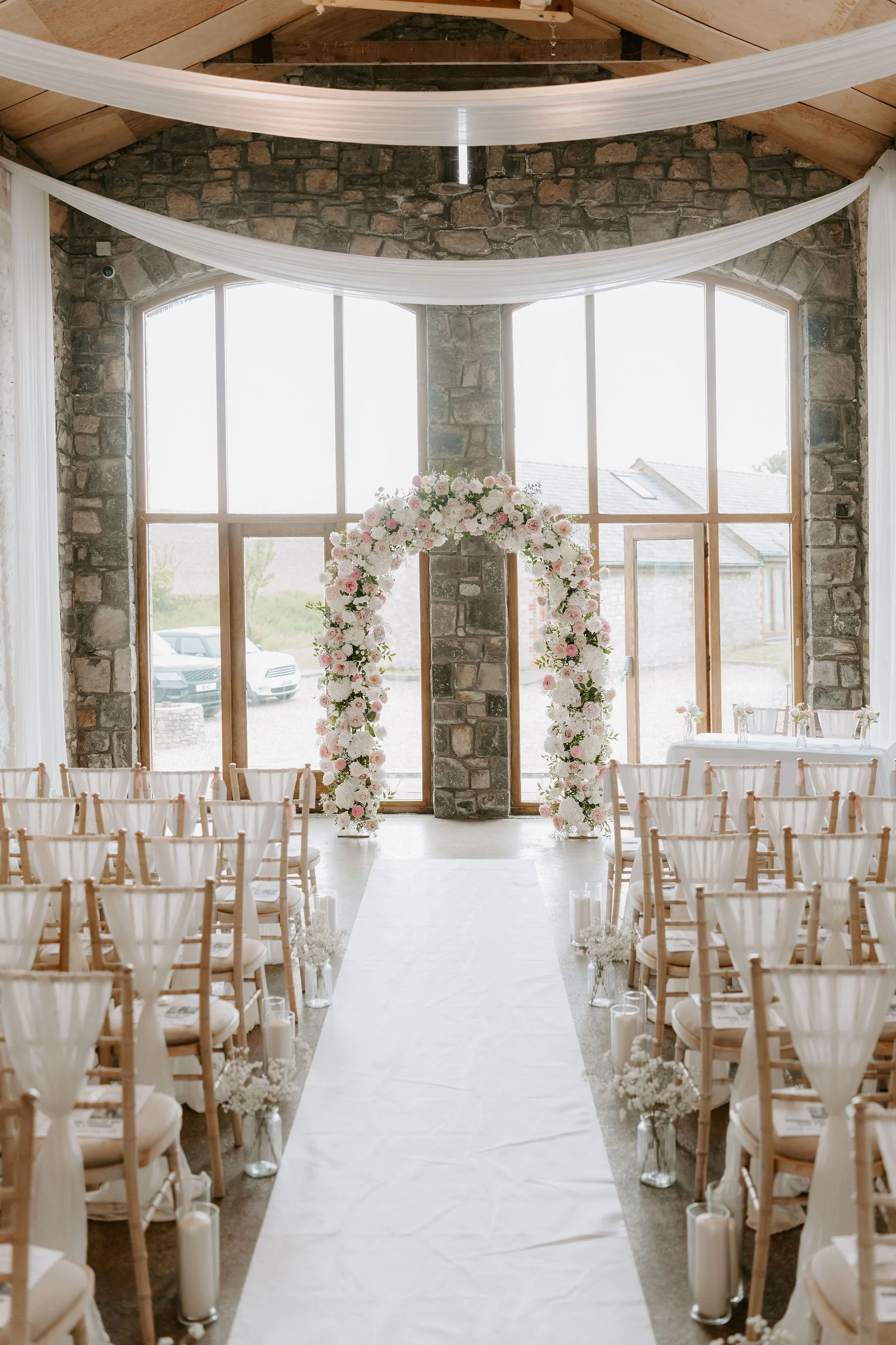 A bright stone-walled ceremony room with white fabric draped below a timber roof, rows of chairs tied with white sashes either side of a white aisle runner, and a round floral arch in front of tall arched windows.