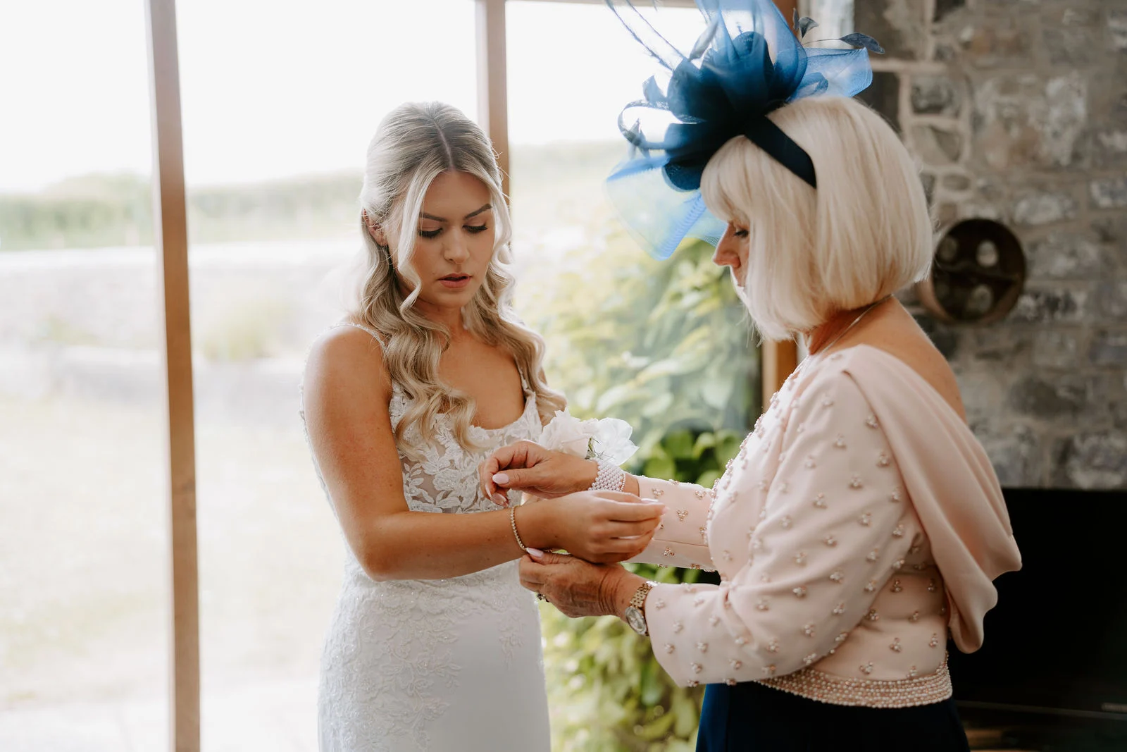 A bride in a lace dress with her hair in loose waves looks down as an older woman in a pale pink cardigan and a navy fascinator fastens a bracelet on her wrist beside a large window