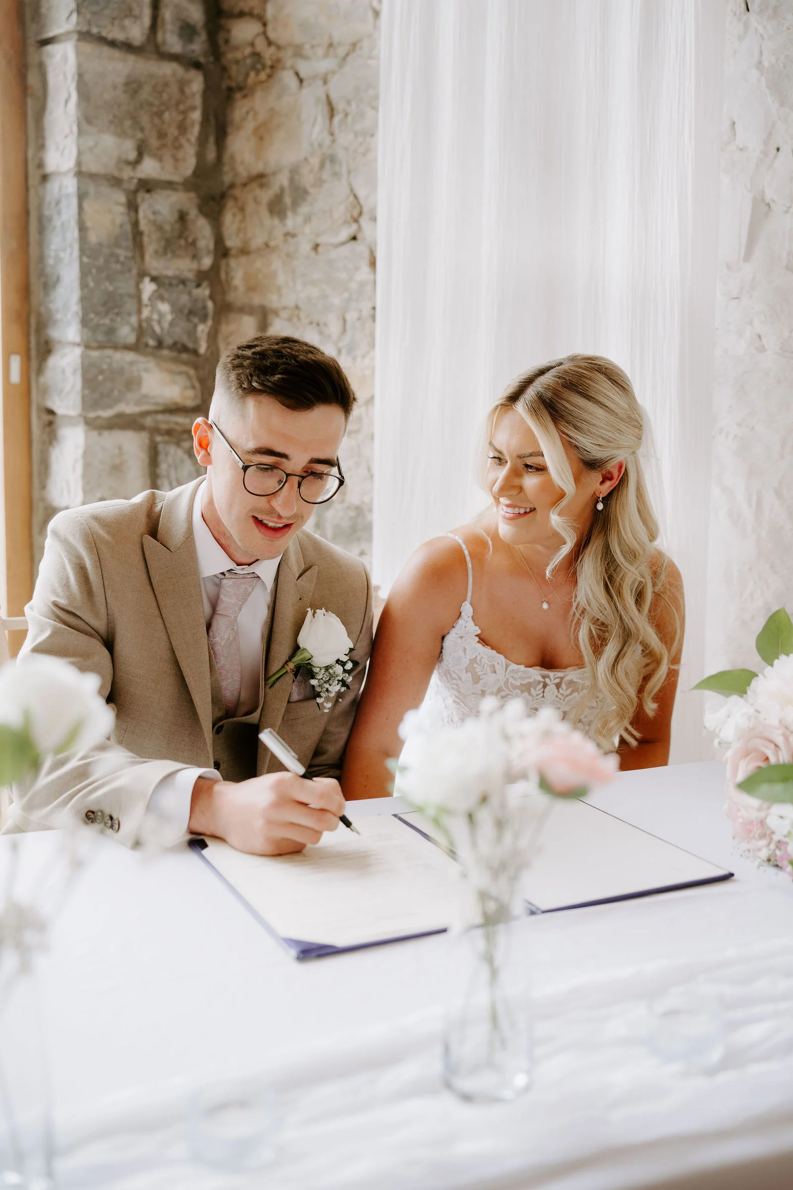 A groom in glasses and a beige suit signs the register while his bride smiles beside him, with a stone wall, white drapes and small vases of white flowers around them