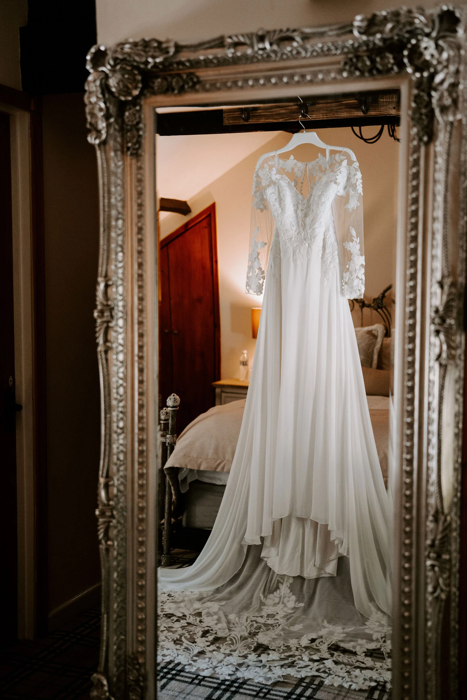 A lace wedding dress with long sleeves hangs in a tall ornate silver mirror in a bedroom, with a tartan carpet on the floor below.