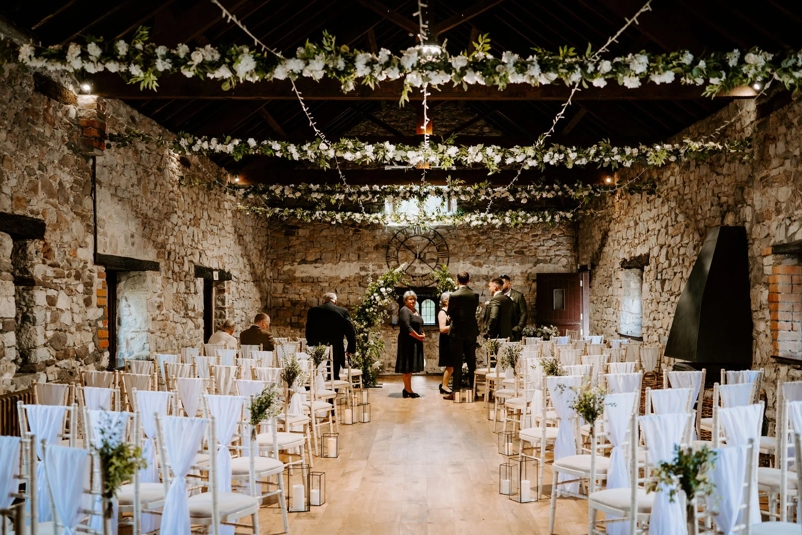 A stone barn set out for a wedding ceremony, with rows of white chairs tied with chiffon, glass lanterns along the aisle and garlands of white flowers and fairy lights hung from the dark timber roof.