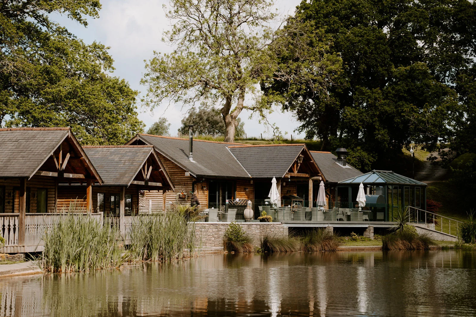Timber lodges with slate roofs and a covered terrace with parasols stand beside a still lake, with tall reeds in front and mature trees behind.
