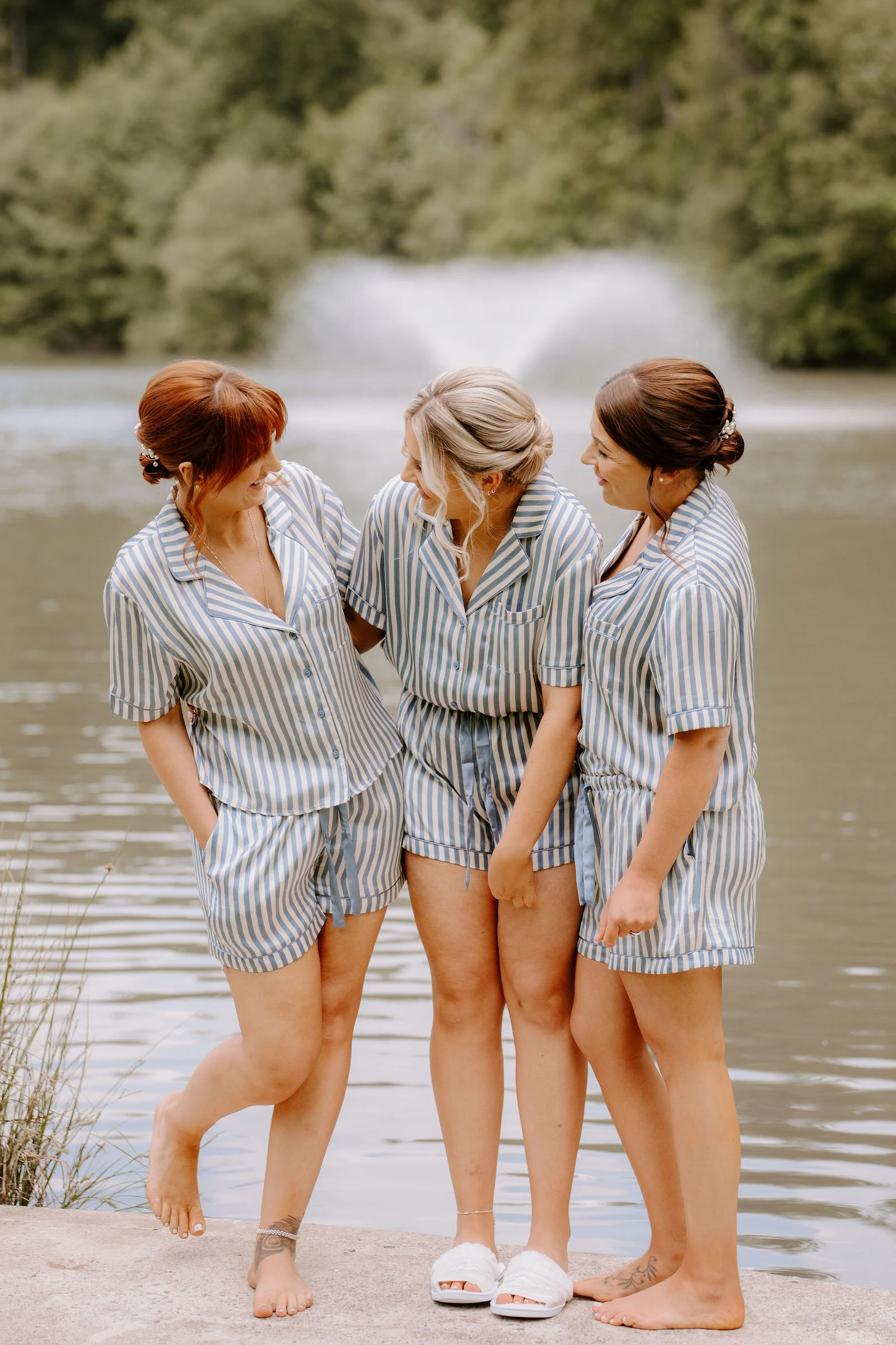 Three women in matching blue striped pyjamas stand arm in arm on a stone ledge beside a lake, laughing at each other, with a fountain and wooded hillside behind them.