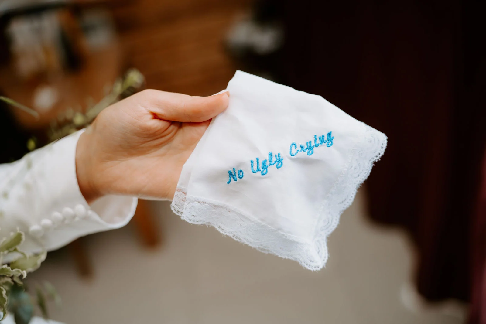 A hand holds up a white lace-edged handkerchief embroidered in blue with the words No Ugly Crying, against a soft dark red curtain.