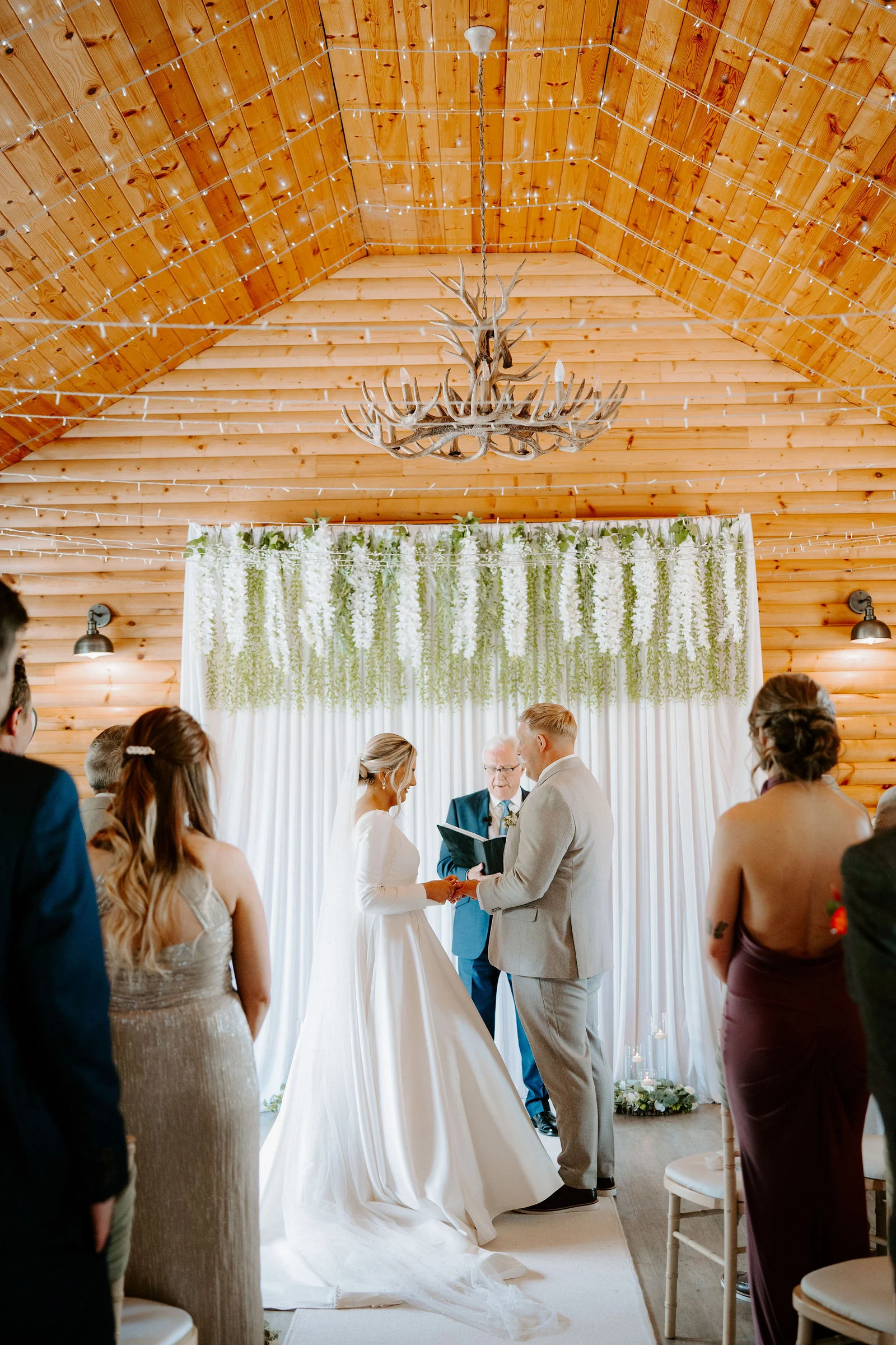 A bride in a long-sleeved white gown and veil and a groom in a light grey suit hold hands while a celebrant in a teal jacket reads from a black book, in front of a white curtain hung with white and green wisteria.