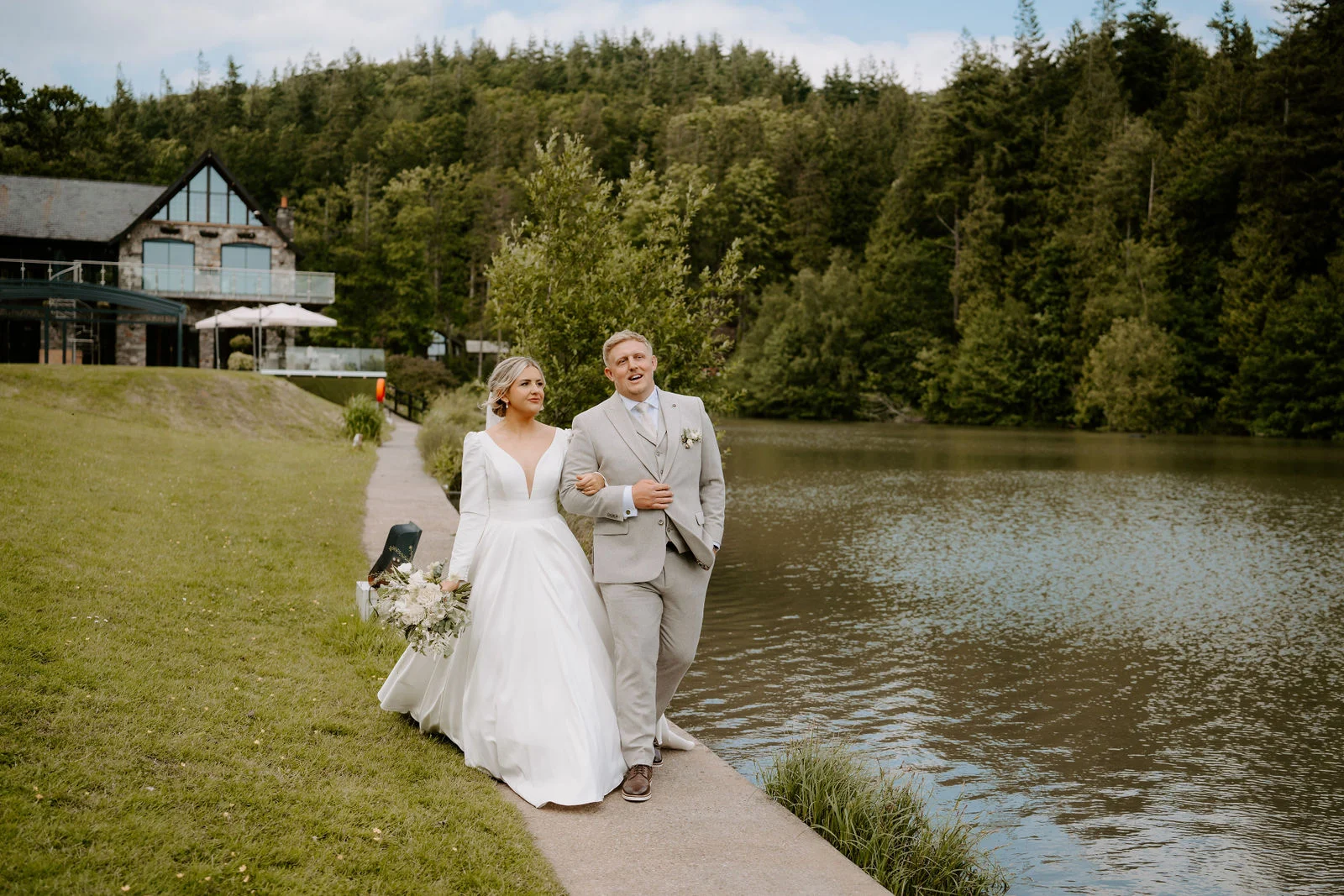 A bride in a long white gown carrying a bouquet and a groom in a light grey suit walk arm in arm along a narrow path beside a lake, smiling at each other, with a stone and glass building and a forested hillside behind.