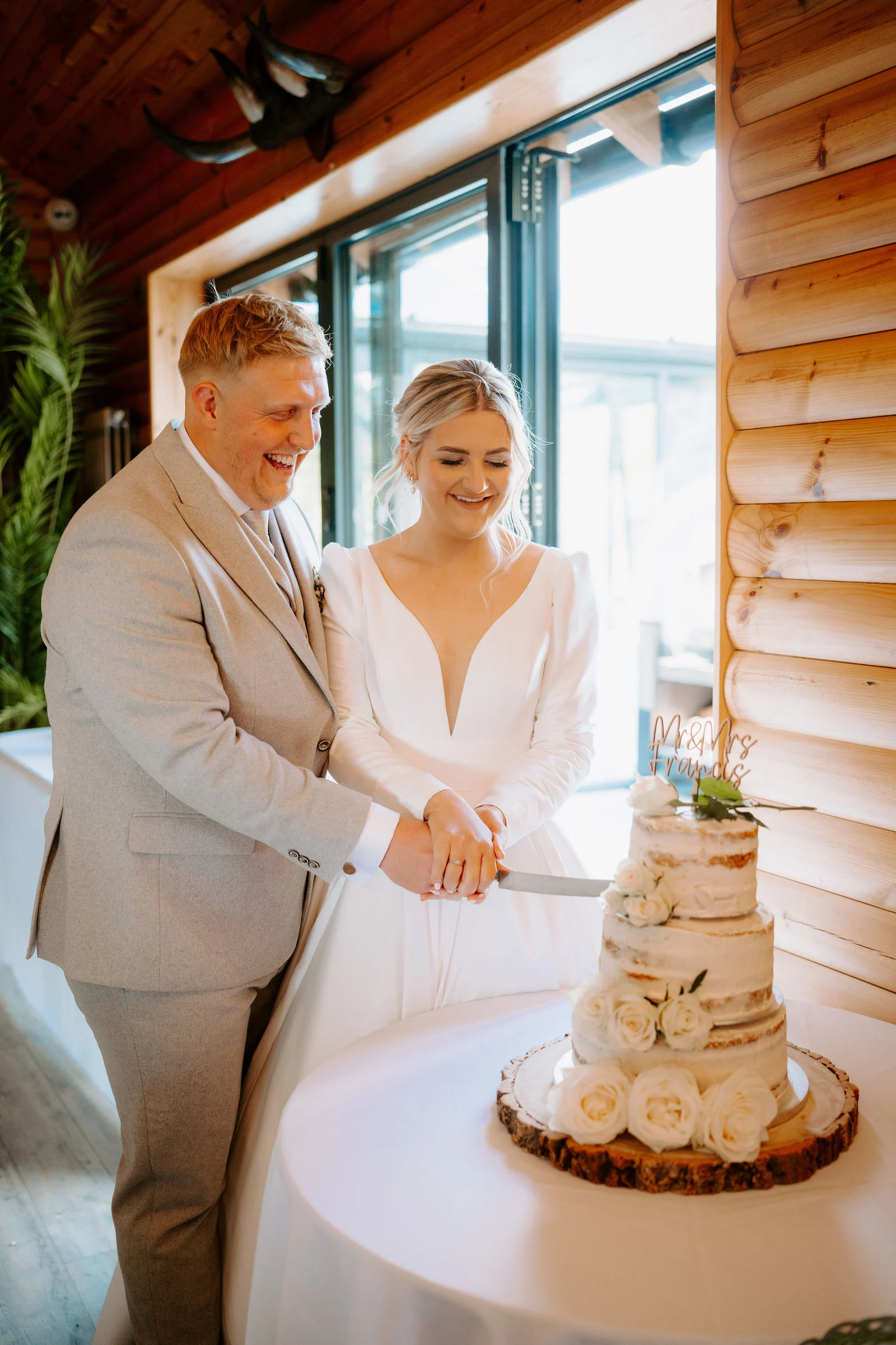 A groom in a light grey suit and a bride in a long-sleeved white gown smile as they cut a tiered naked cake decorated with white roses, beside a log wall and a large window.
