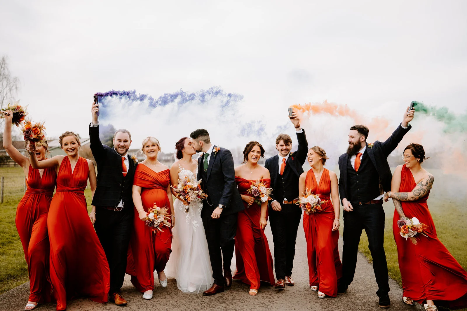 The wedding party walks along a path towards the camera, laughing and holding up blue, orange and green smoke flares, the bridesmaids in rust-red dresses and the groomsmen in dark suits, with the couple kissing in the middle.