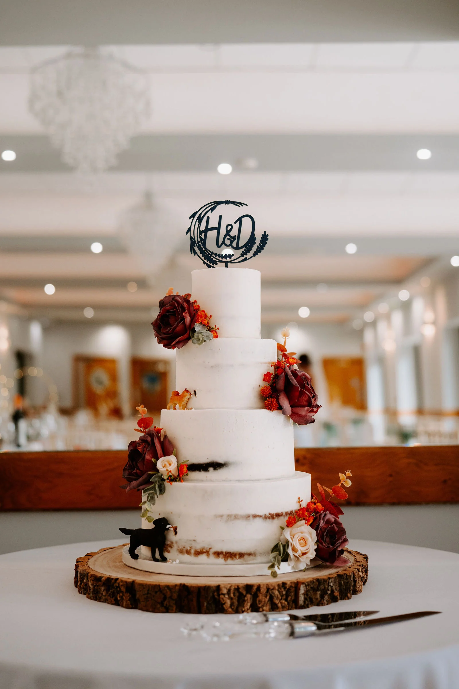 A tall, lightly iced wedding cake on a wooden log slice, decorated with dark red roses, autumn leaves and a small black dog figure, with a round topper on top and a softly blurred reception room behind.