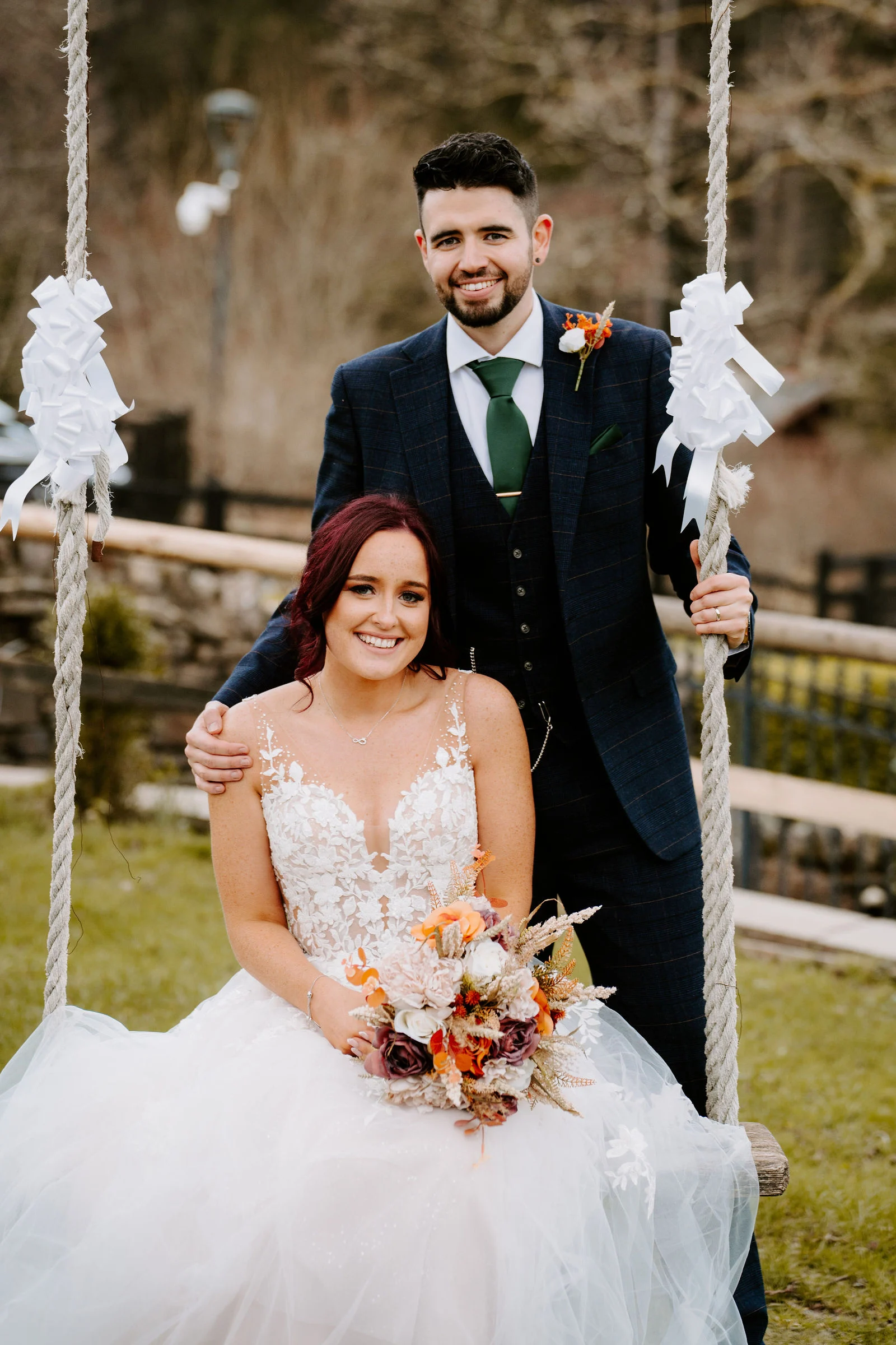 A bride in a white lace gown sits on a rope swing tied with white bows and holds an autumn-coloured bouquet while the groom in a navy checked suit stands behind her with a hand on her shoulder, both smiling, with a stone wall and timber fence behind