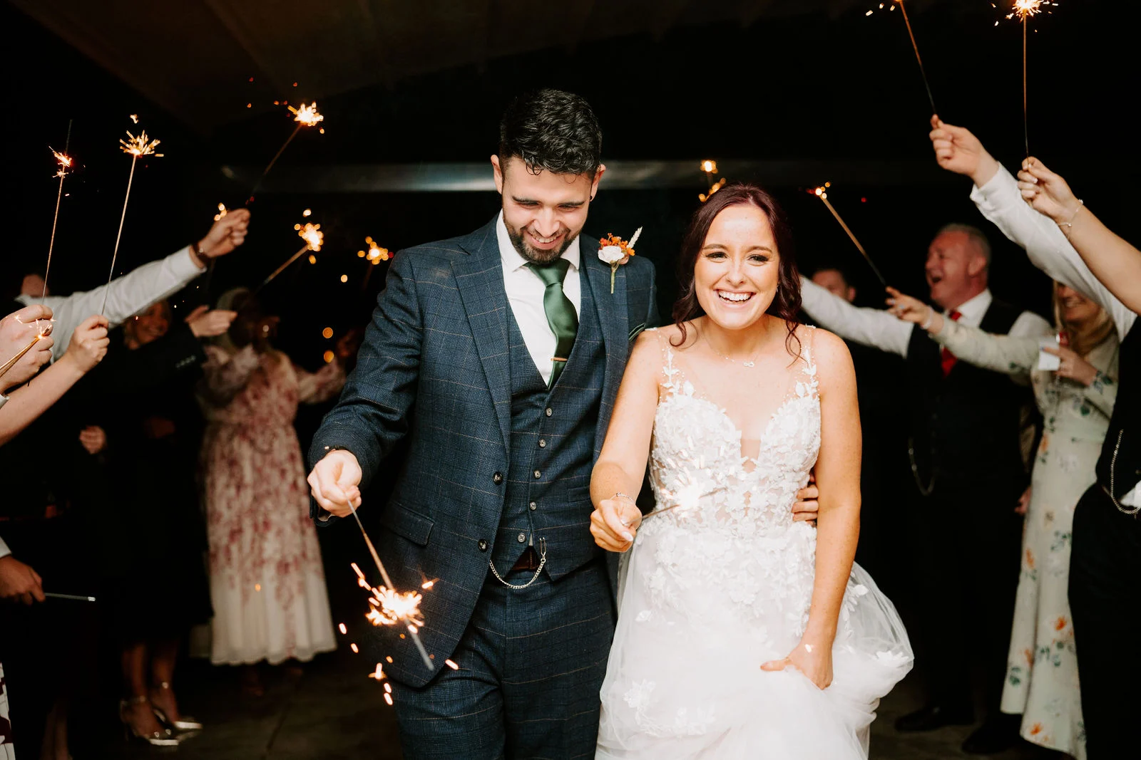 A laughing bride and groom walk arm in arm through a tunnel of guests holding lit sparklers at night, the groom holding a sparkler low in one hand.