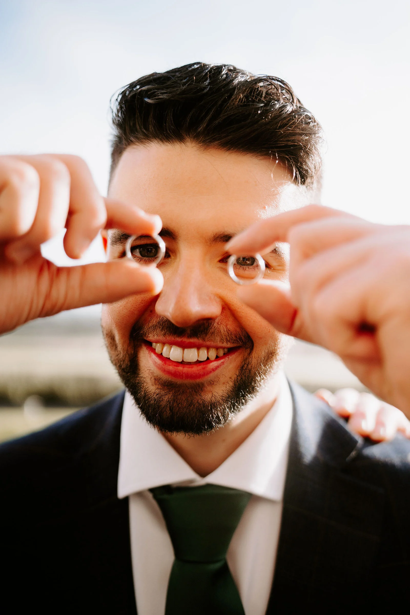 A smiling groom in a dark suit and green tie holds two wedding rings up to his eyes like glasses, with open countryside softly blurred behind him.