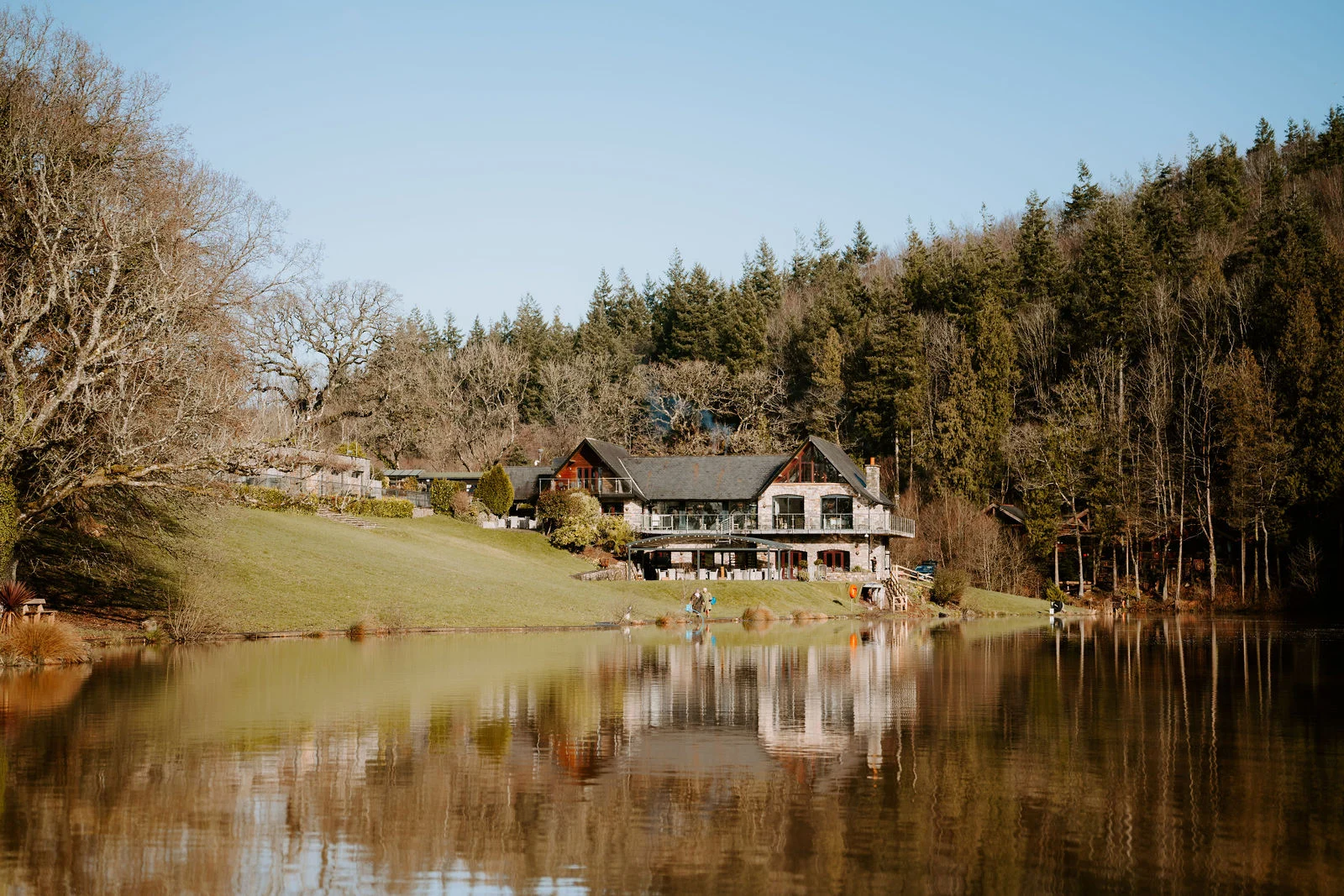 A large stone and timber building stands on a grassy bank across a still lake, reflected in the water, with wooded hillsides behind it under a clear blue sky.