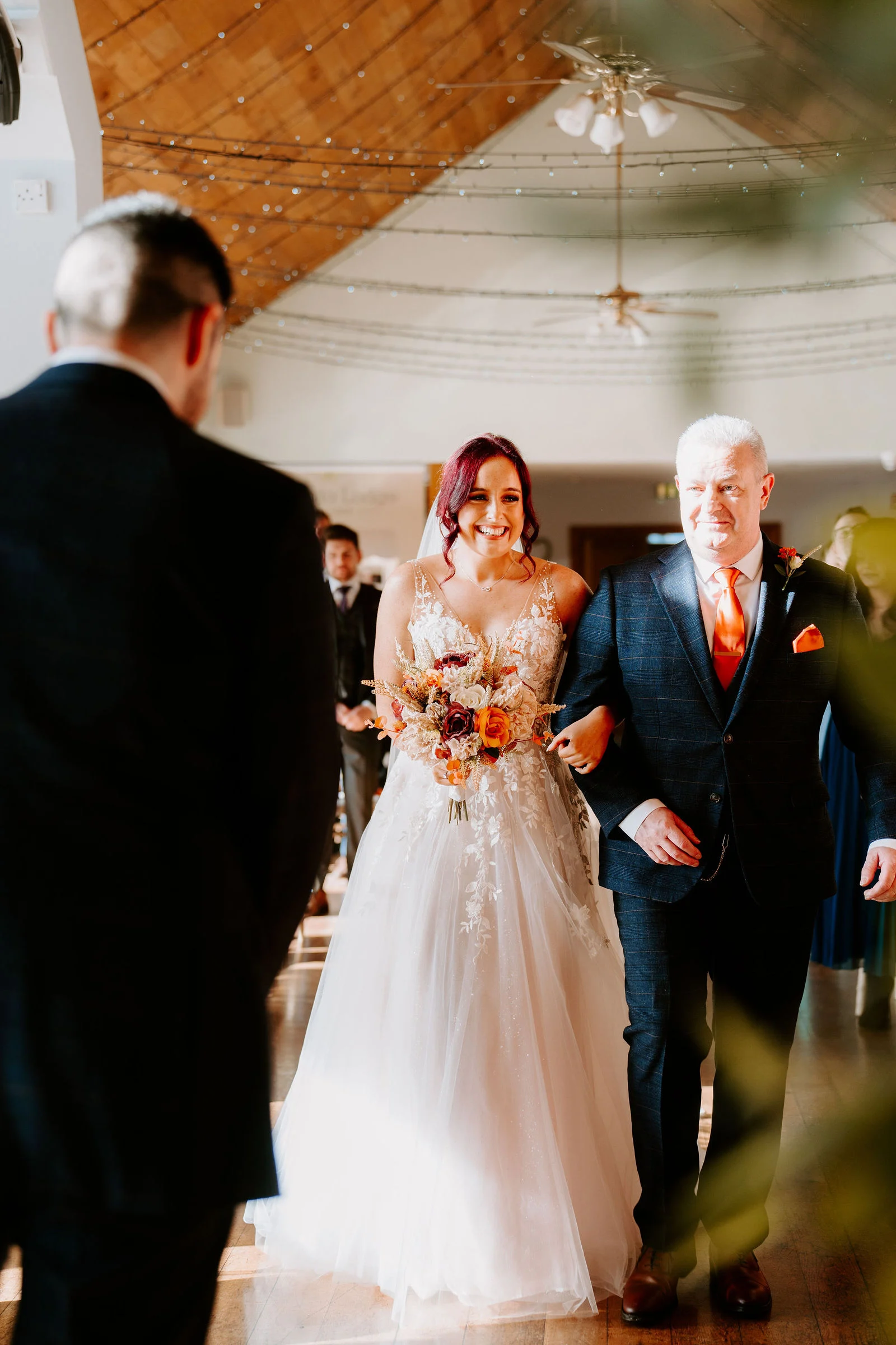 A smiling bride in a lace-bodice gown carries an orange and cream bouquet down the aisle on the arm of a man in a blue checked suit, under a timber-beamed ceiling.